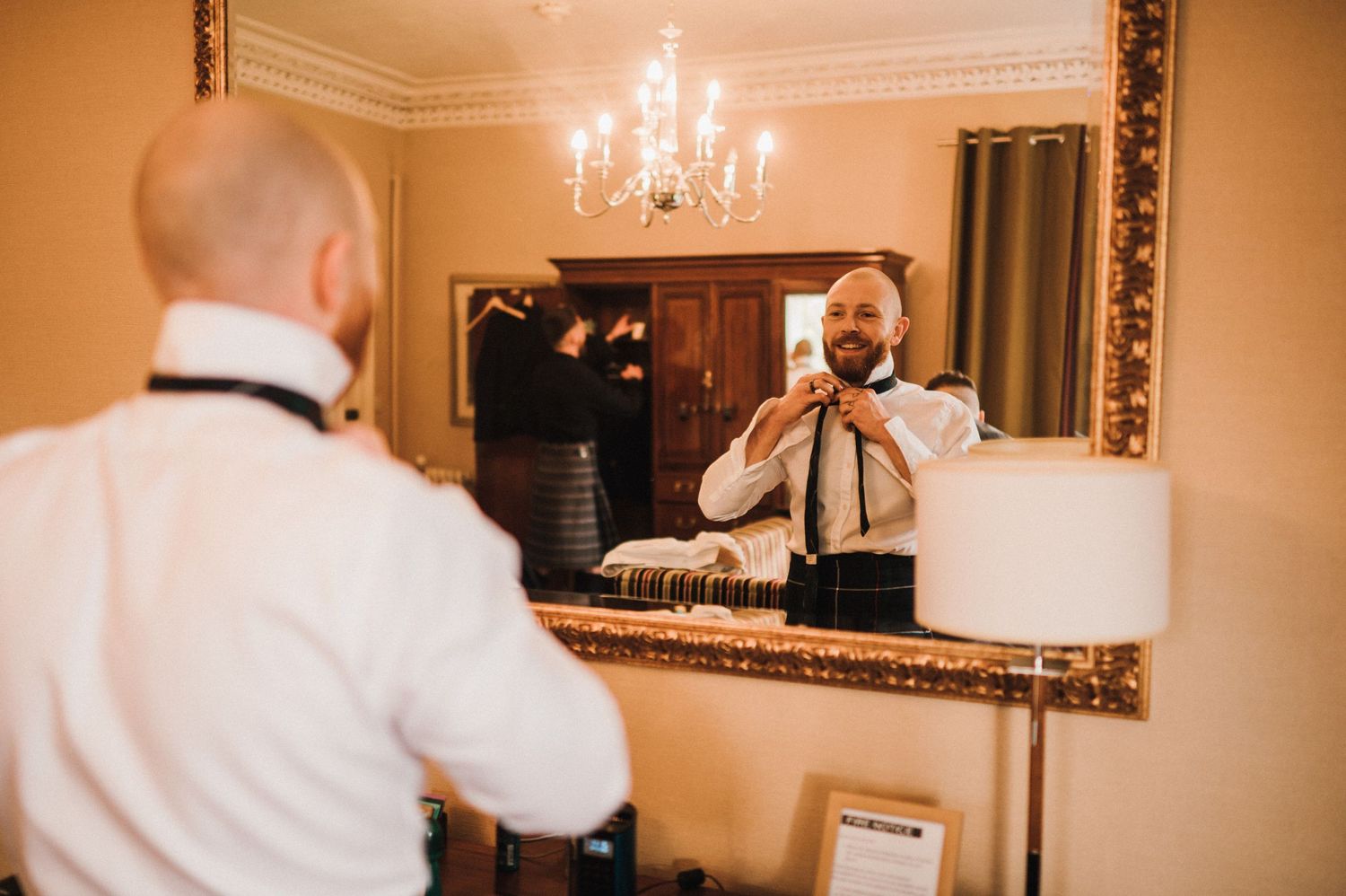 Reflection in a gilded mirror shows someone adjusting a tie while getting dressed in a formal setting.