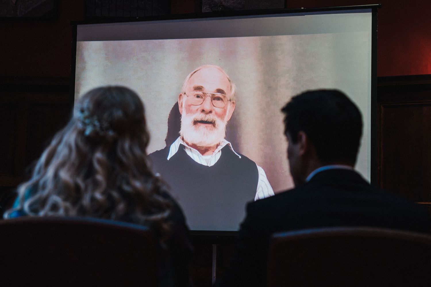 Two silhouettes watch a projected video presentation featuring an elderly speaker.