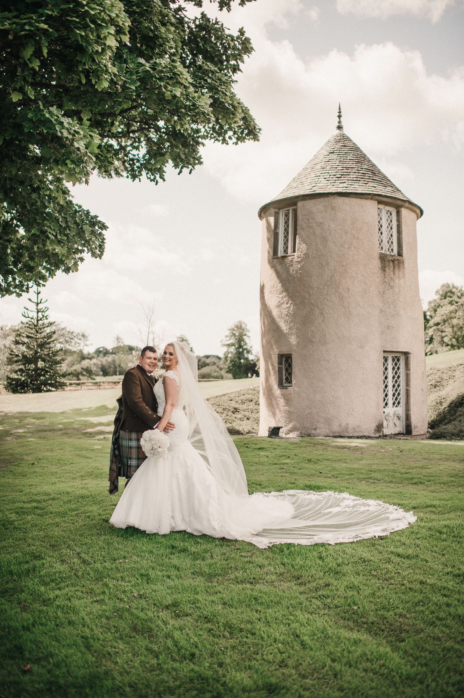 A wedding couple poses in front of a round stone tower on a lush green lawn with trees in the background.