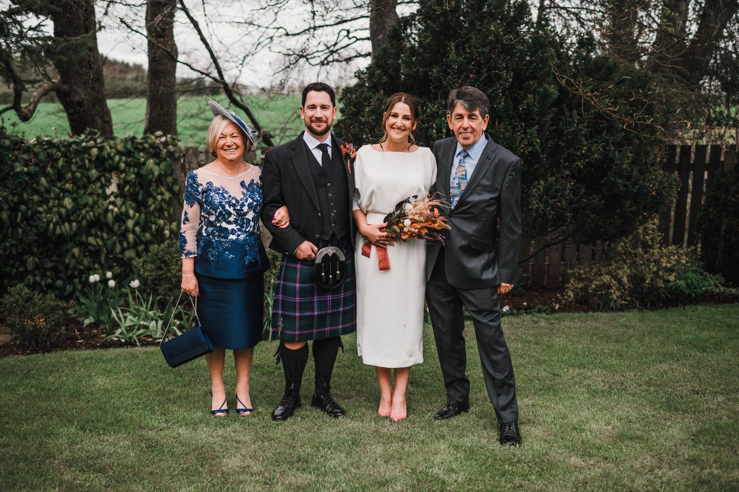 Wedding party in a garden setting with traditional Scottish kilt and fall-themed bouquet.