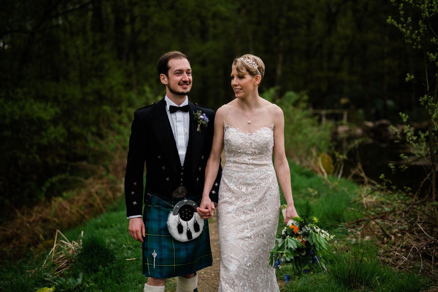 A couple in wedding attire pose outdoors, with the groom wearing a traditional Scottish kilt.
