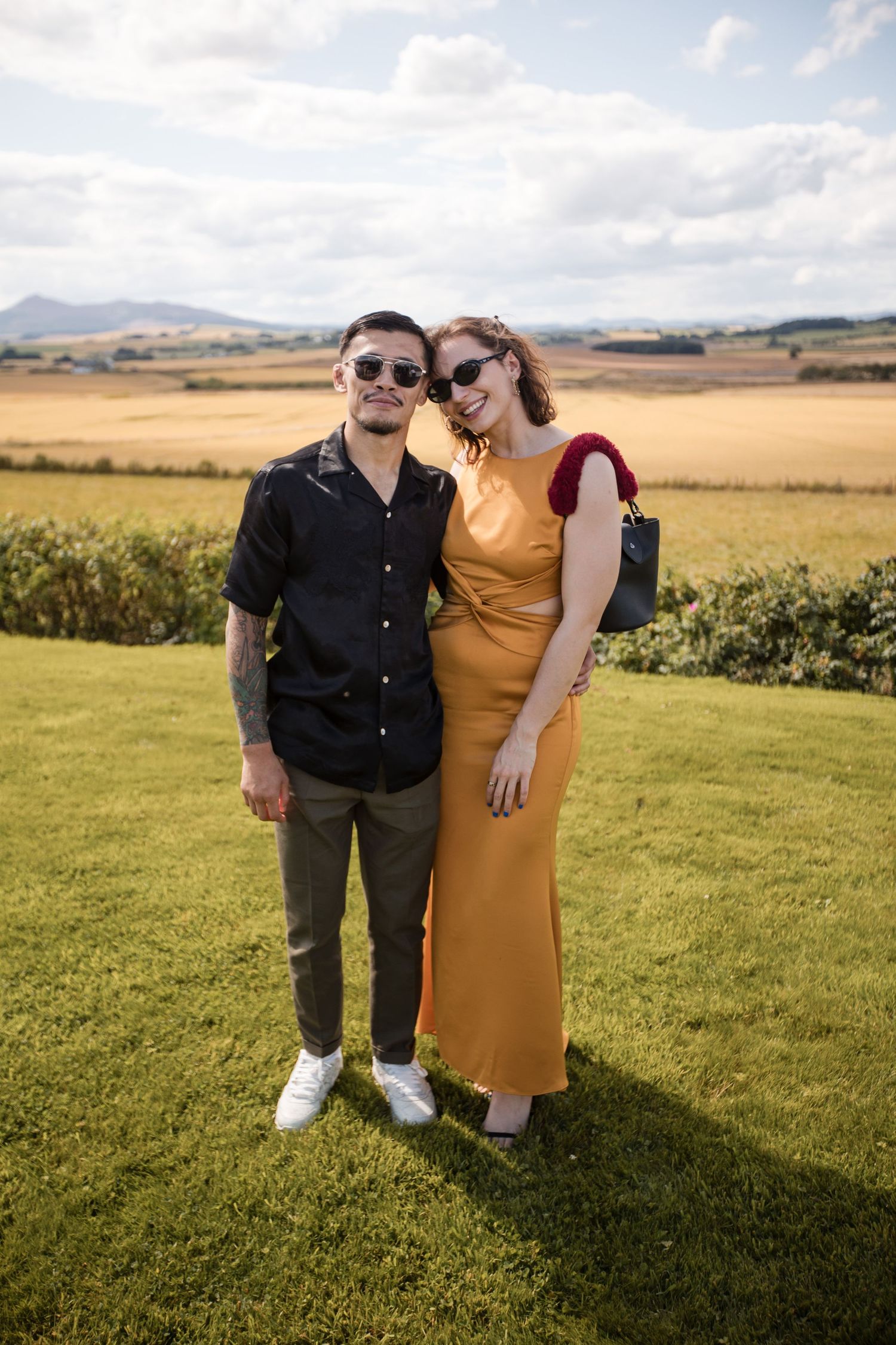 Couple poses together on green grass with scenic countryside and mountains in background.