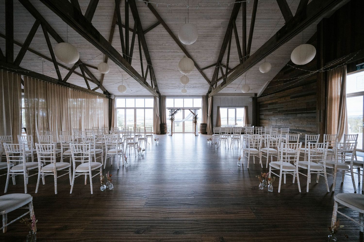 Spacious rustic venue interior with white chairs set up for an event and natural light streaming through windows.
