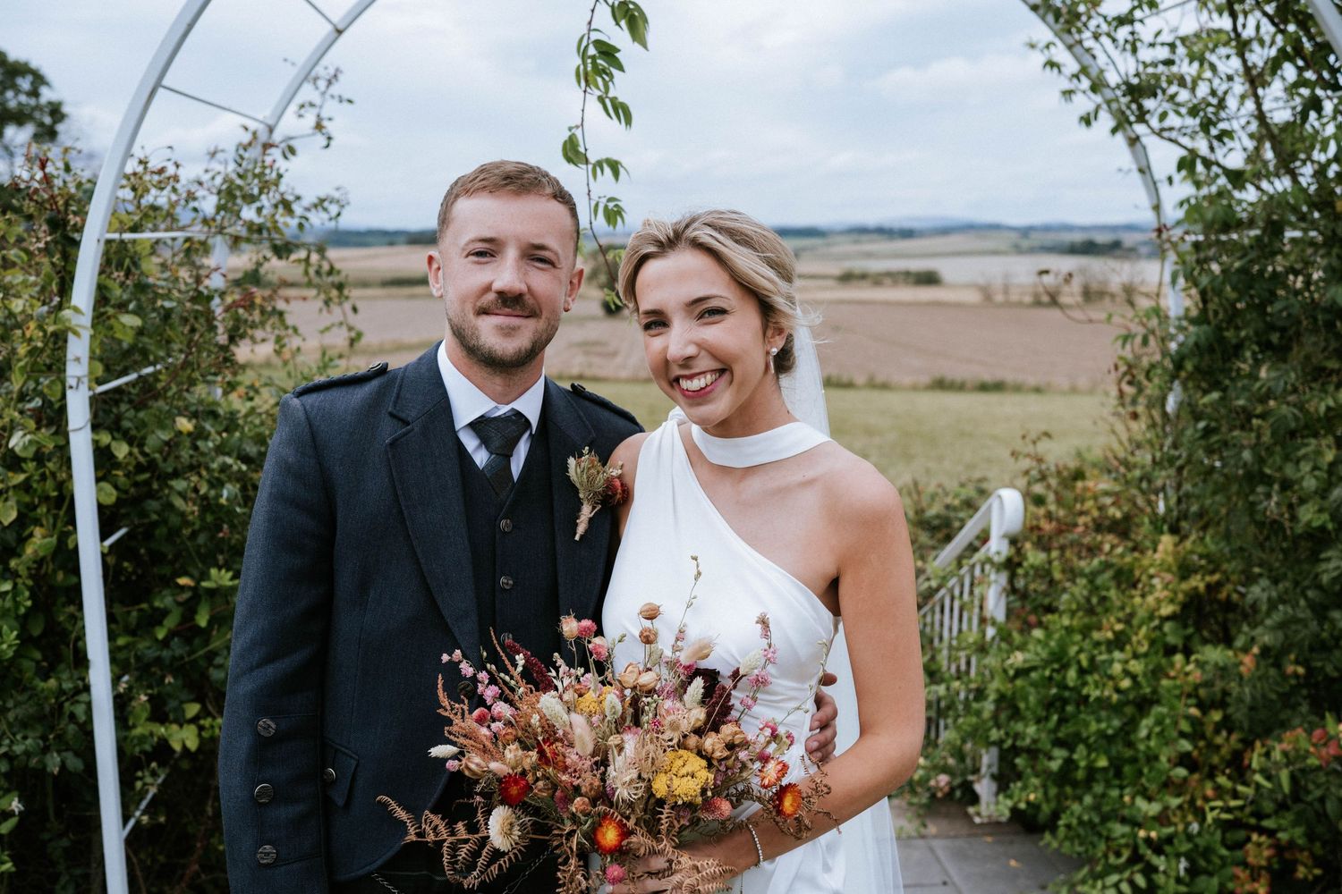 Newlyweds pose under an outdoor arch with dried flower bouquet against countryside backdrop.