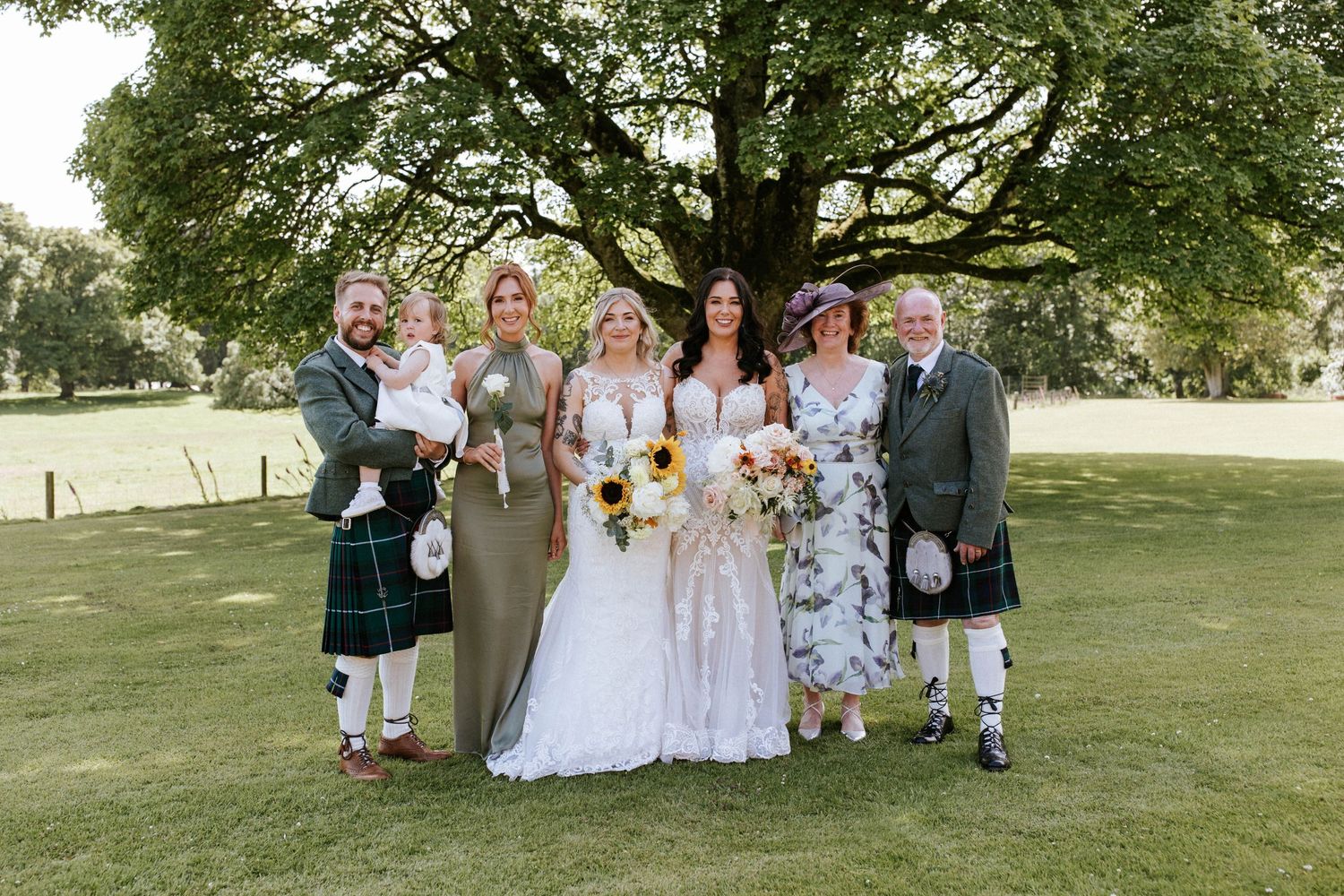 Wedding celebration group poses together on grassy hill with traditional Scottish attire.