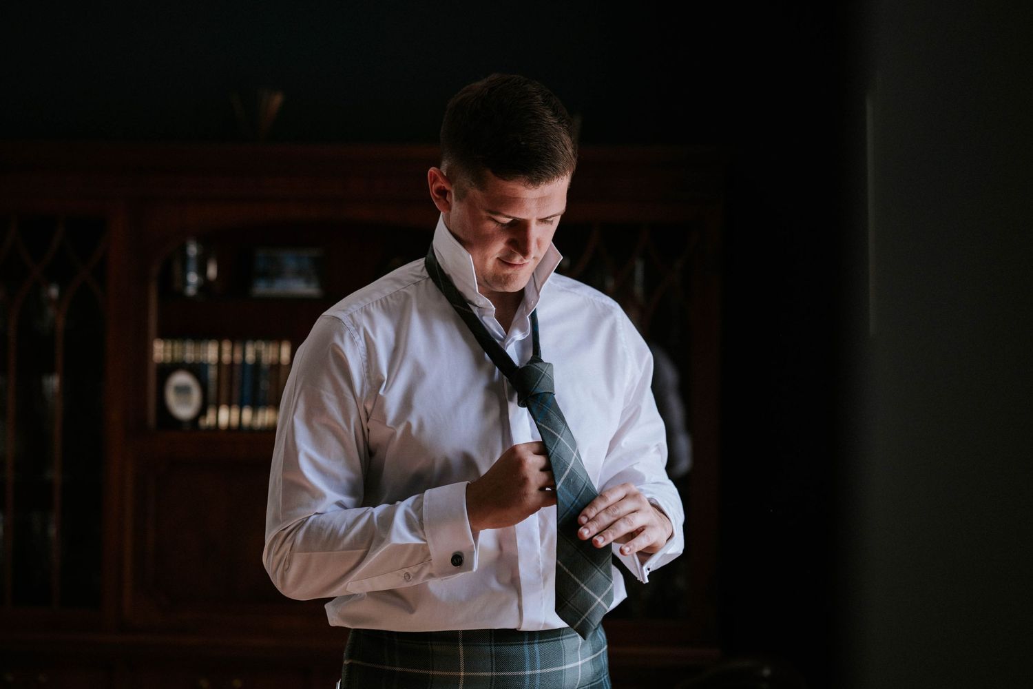 Groom getting ready in formal Scottish attire in dimly lit room.
