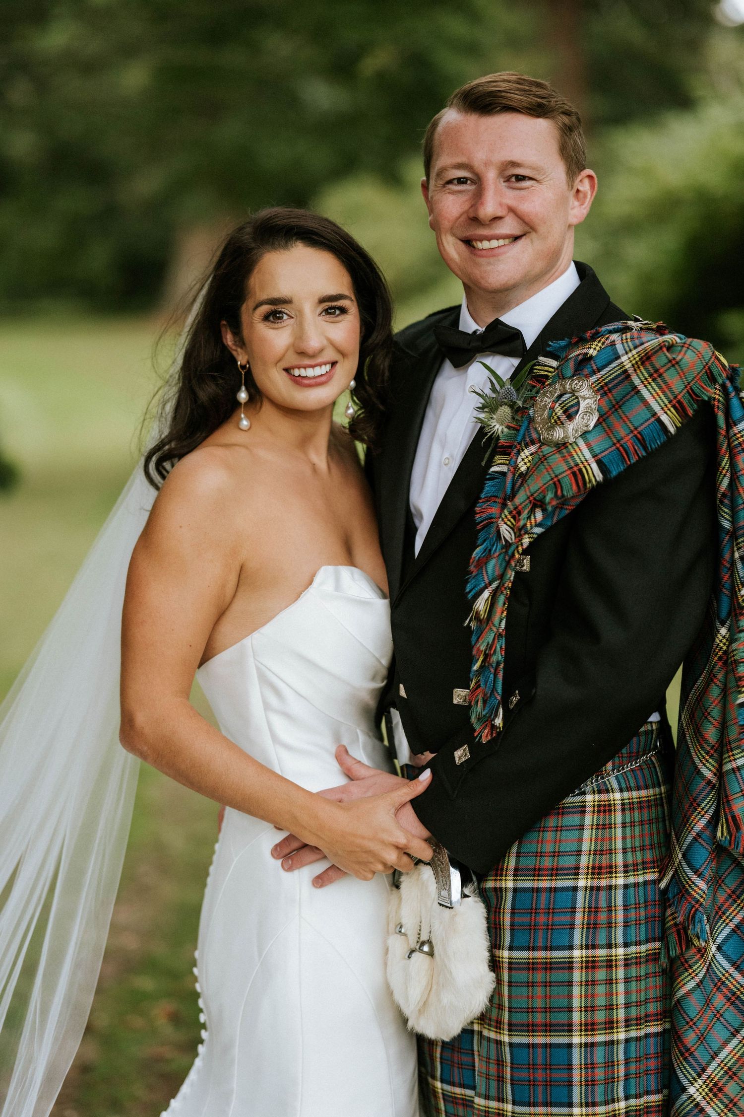 Newlyweds pose together outdoors, with groom wearing traditional Scottish kilt and bride in white gown.
