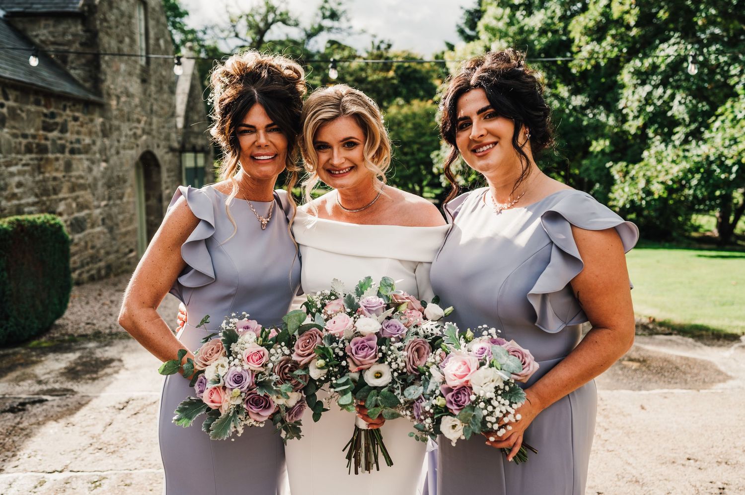 Bridesmaids in lavender dresses holding pink and white floral bouquets outside stone church.