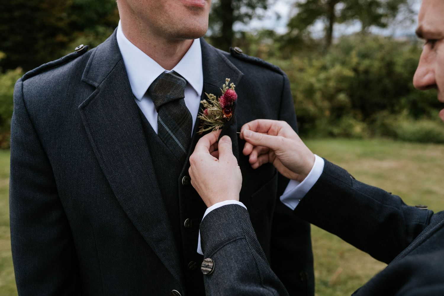 Groomsmen in black suits and kilts adjusting boutonnieres outdoors.