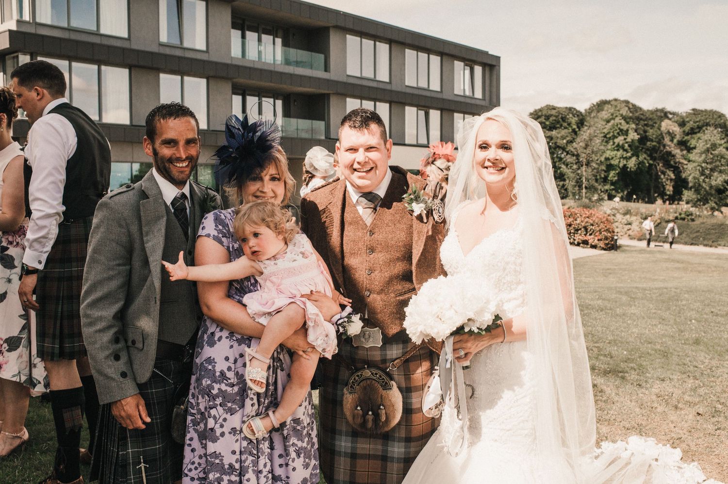 A wedding party gathered outside a modern building on a sunny day.