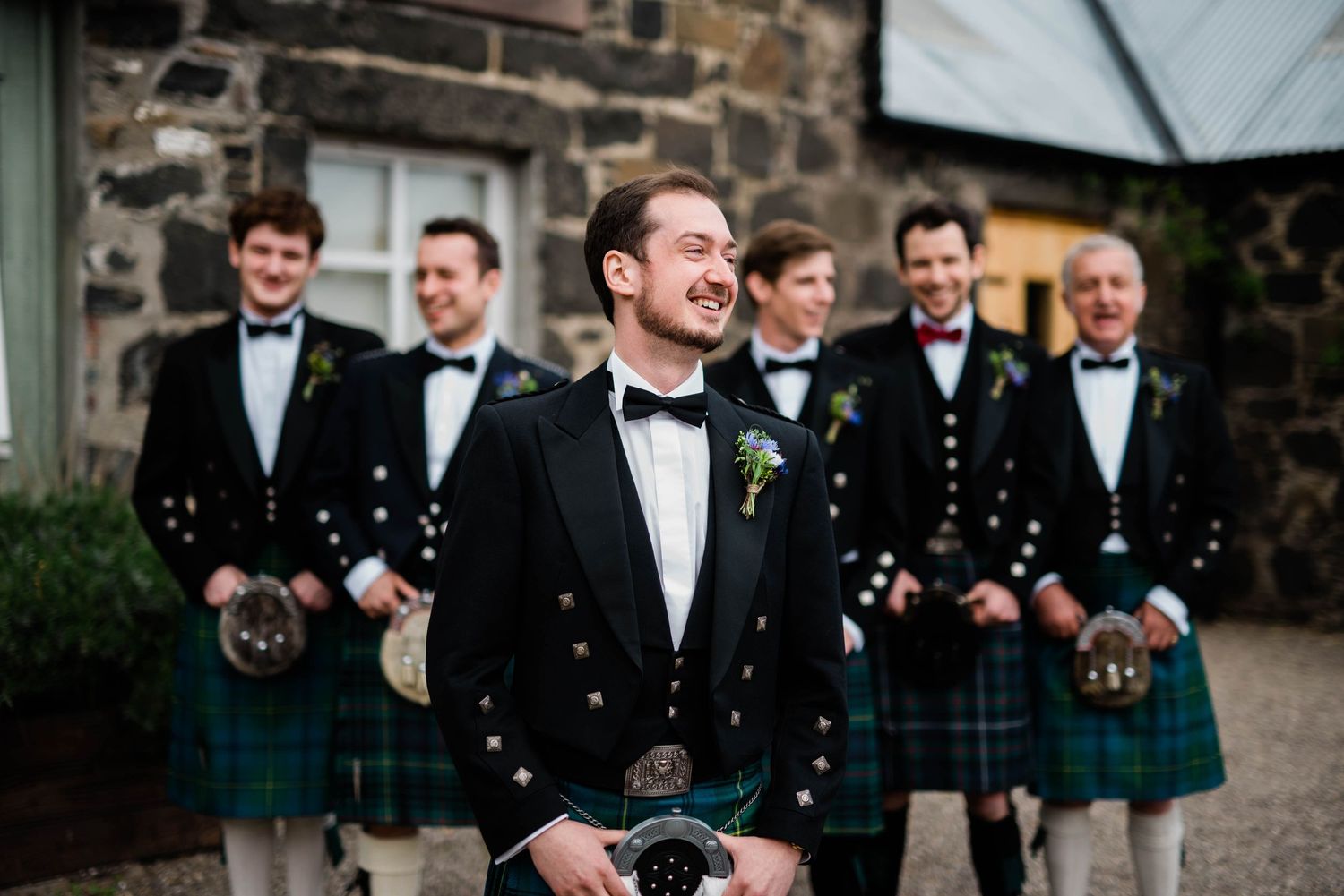 Groomsmen in traditional Scottish kilts stand behind central figure in bowtie.