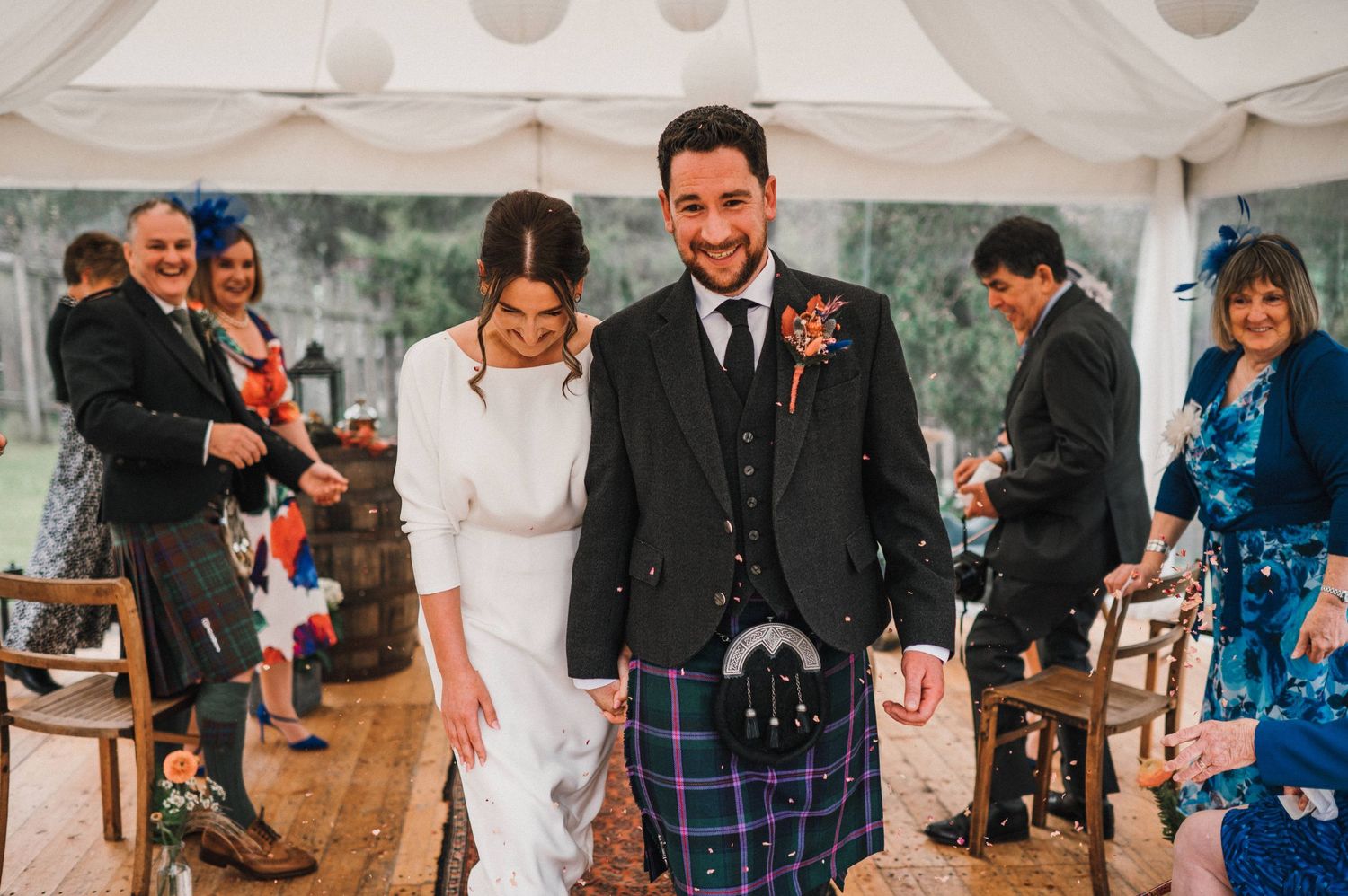 Joyful wedding celebration under white tent with guests applauding in traditional Scottish attire.