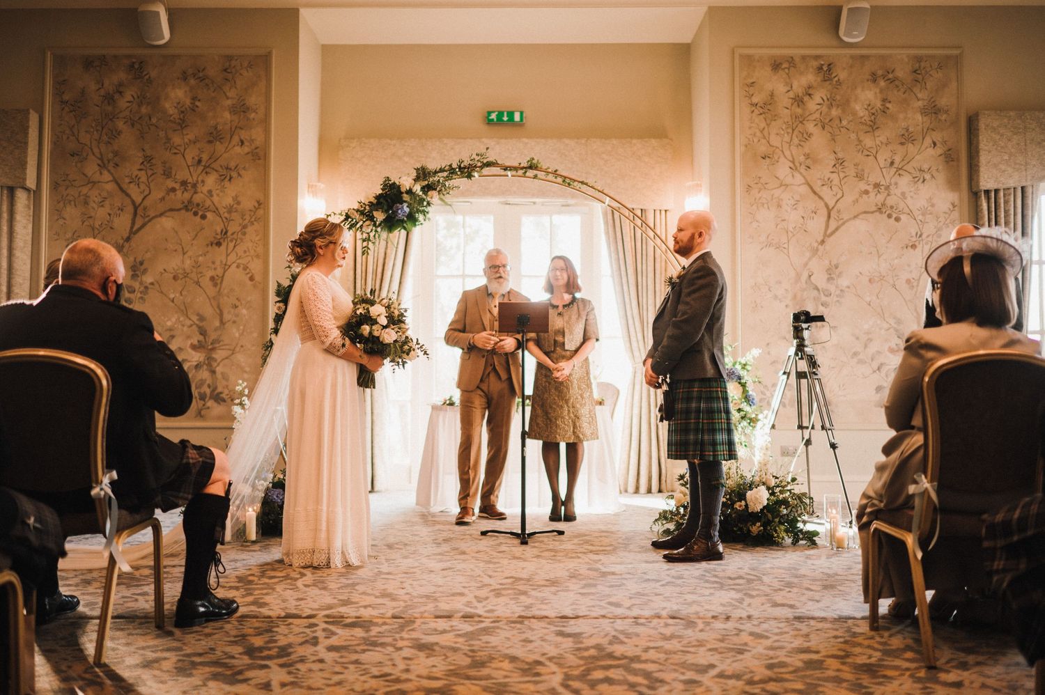 Elegant wedding ceremony in a room with floral wallpaper and an archway decorated with greenery.
