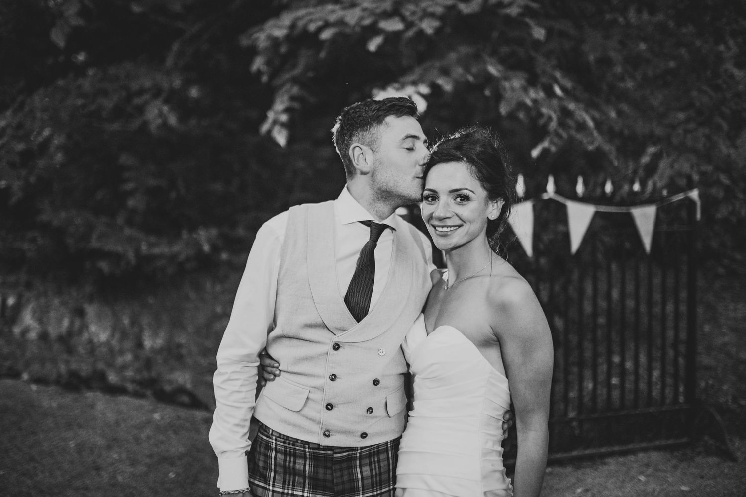 A black and white photo shows a romantic moment between newlyweds in a garden setting.