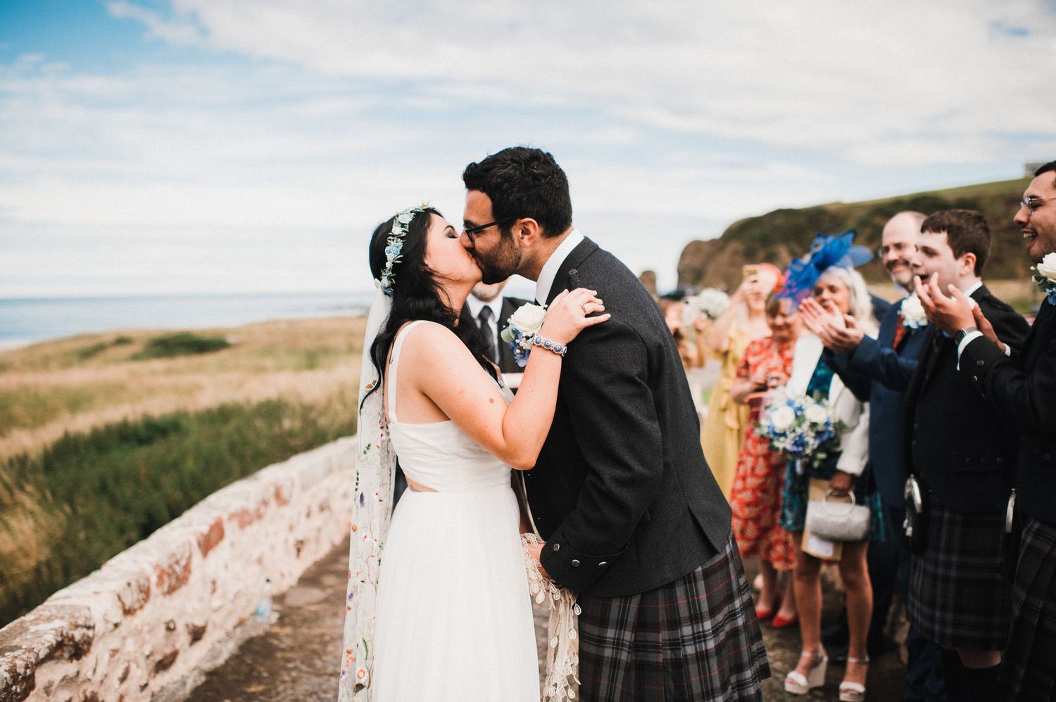Newlyweds share their first kiss surrounded by wedding guests on a scenic hilltop path in Scotland.