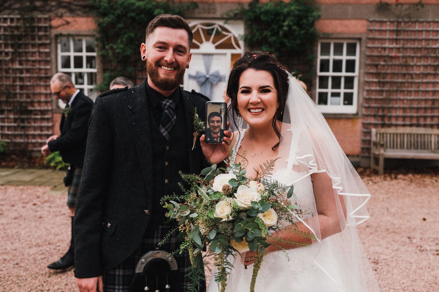 Newlyweds in traditional Scottish wedding attire pose outside brick manor house.