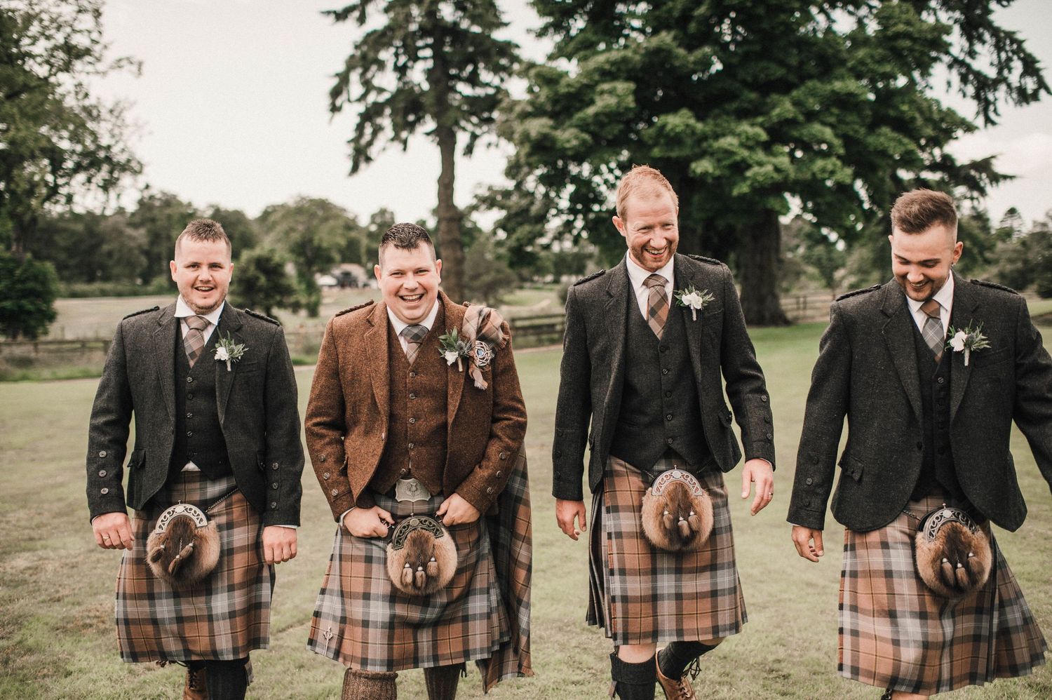 Four people wearing traditional Scottish kilts stand together on grass.