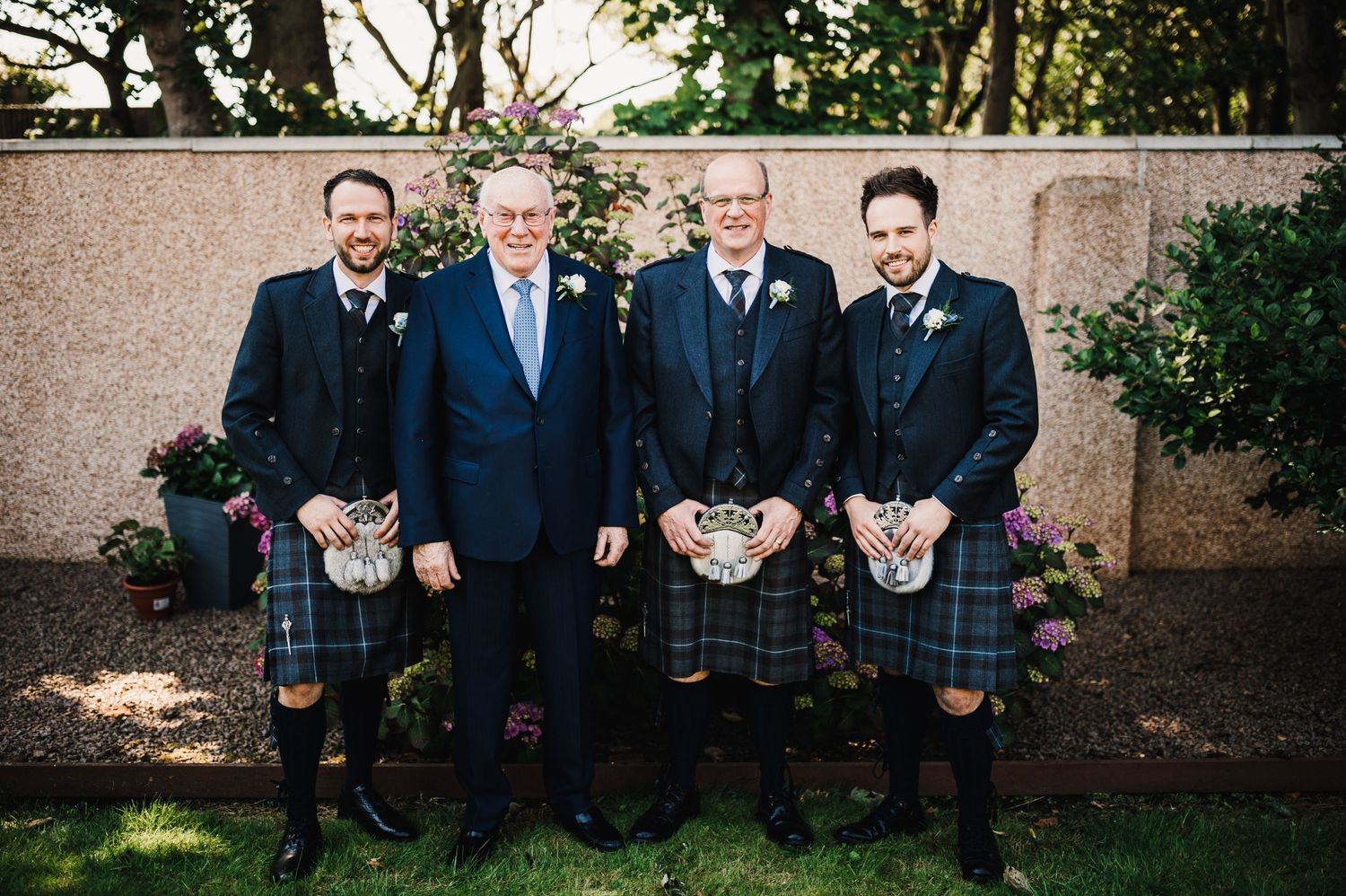 Group portrait of men in formal suits and kilts standing against stone wall.