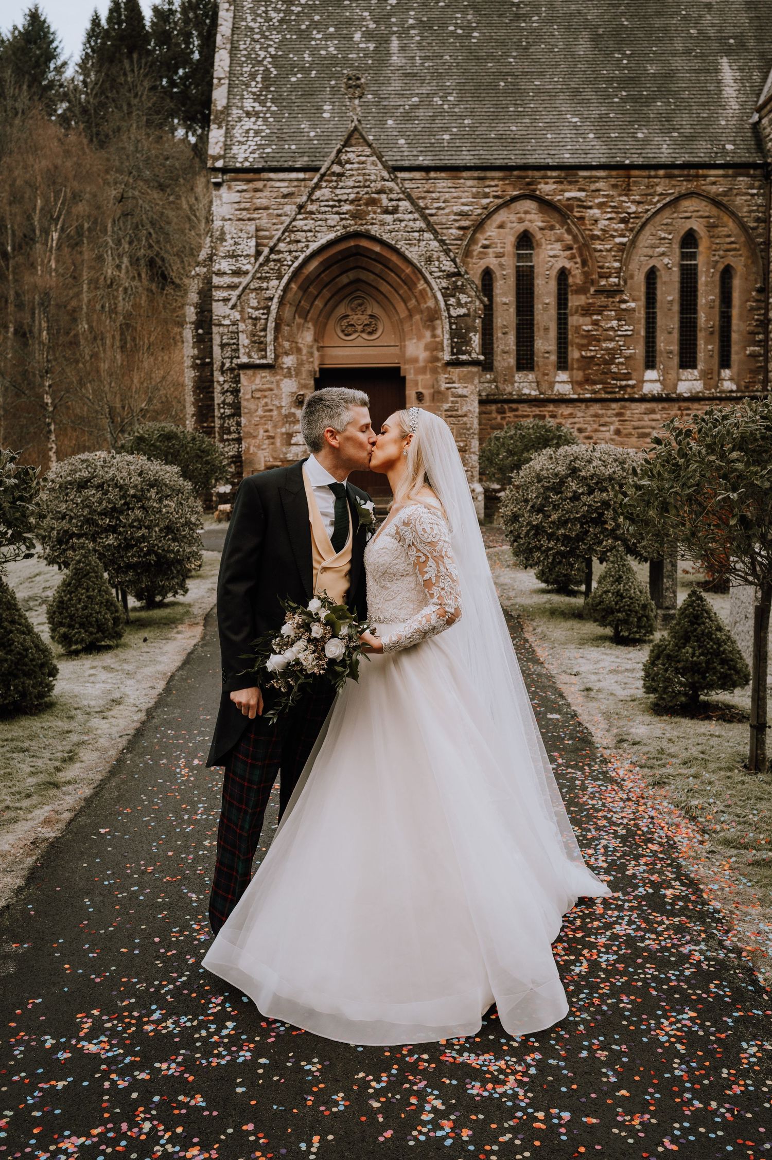 Romantic wedding moment captured outside stone church with gothic architecture and manicured gardens.