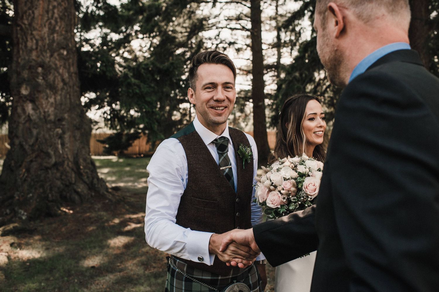 Groom in a vest shakes hands during wedding ceremony in forest setting.