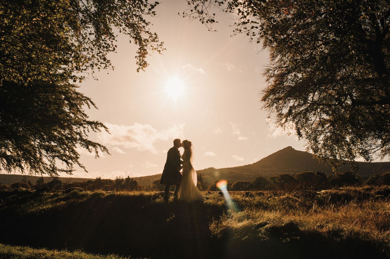Romantic sunset silhouette of newlyweds framed by trees against mountain backdrop.