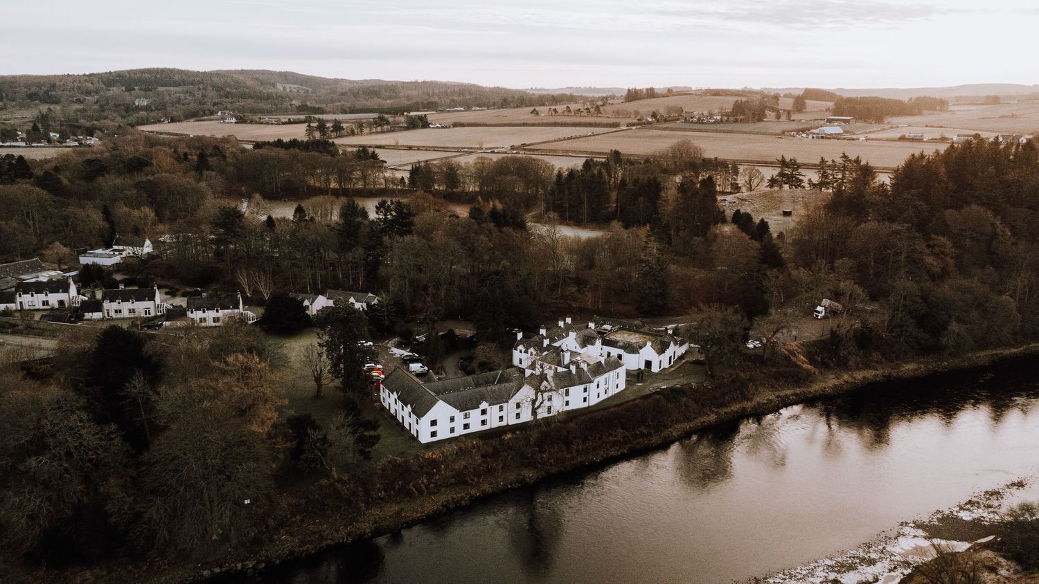Aerial view of white buildings nestled along a river bank in a scenic rural landscape.