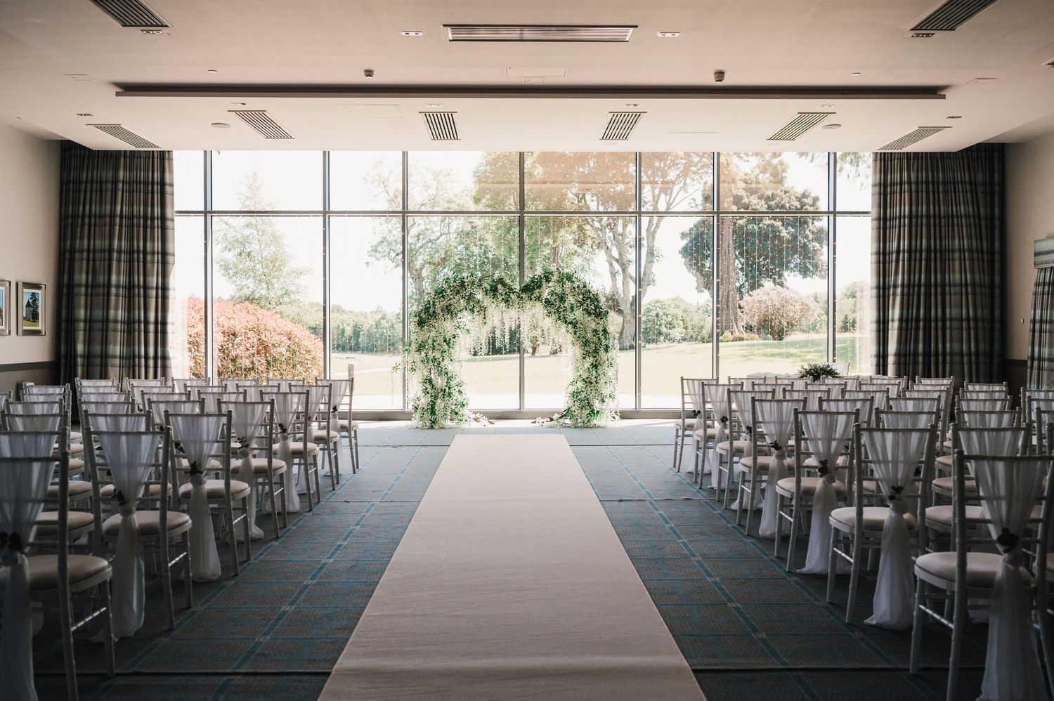 Indoor wedding ceremony space with white aisle runner, grey chairs and floor-to-ceiling windows overlooking nature.