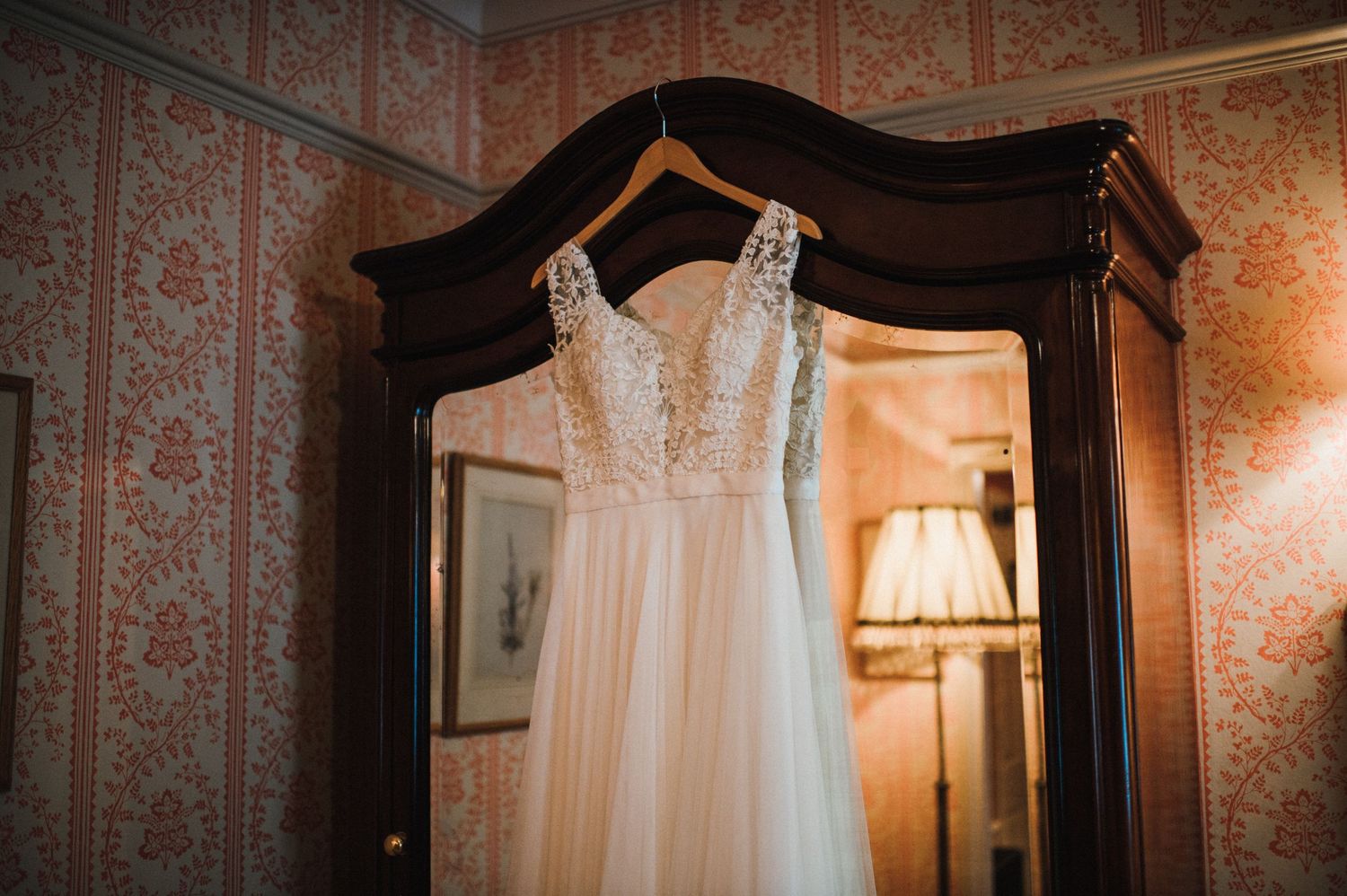A white lace wedding dress hangs on a wooden armoire against floral wallpaper.