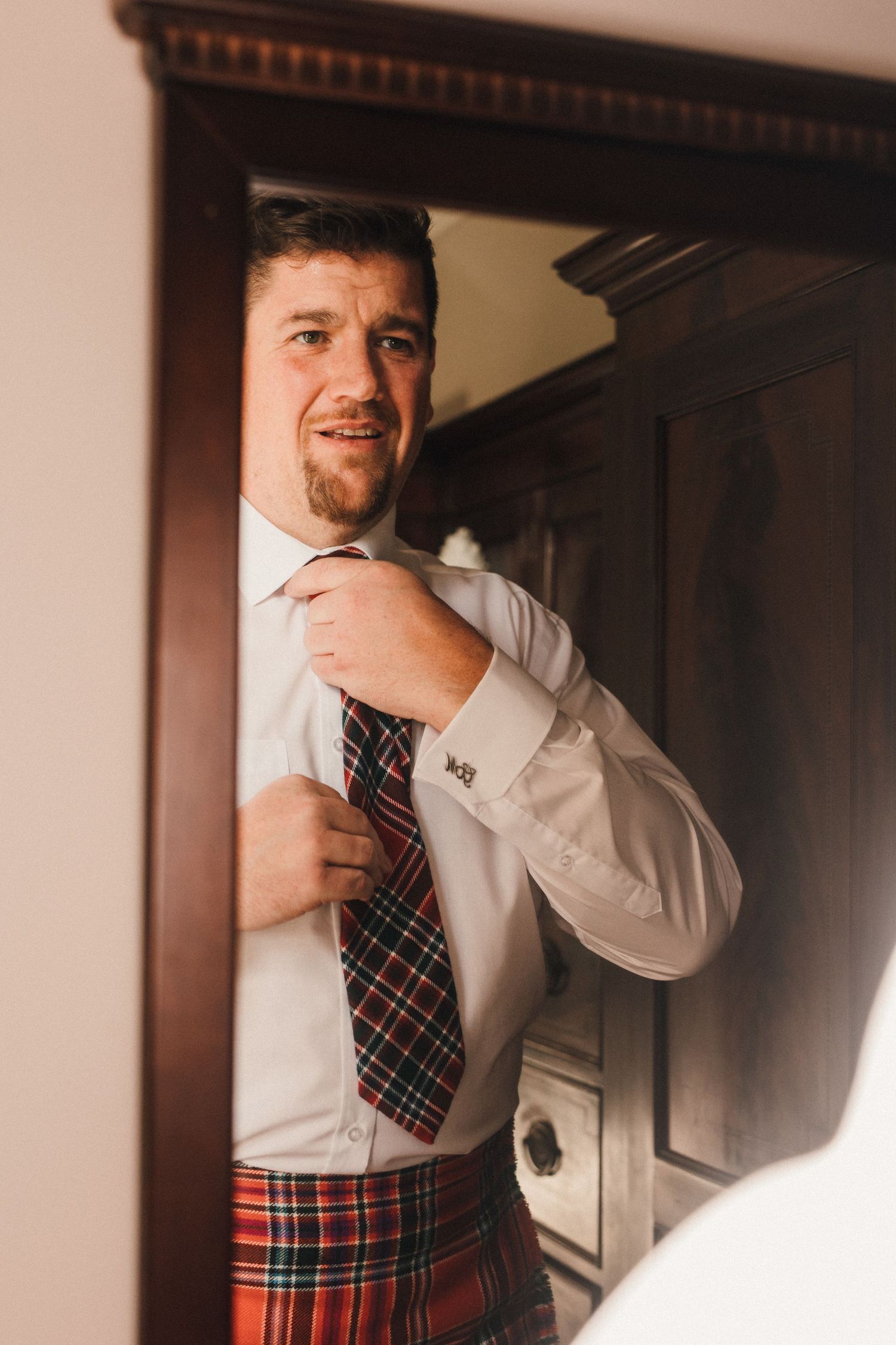 Person wearing white shirt and plaid kilt adjusting tie in mirror reflection.