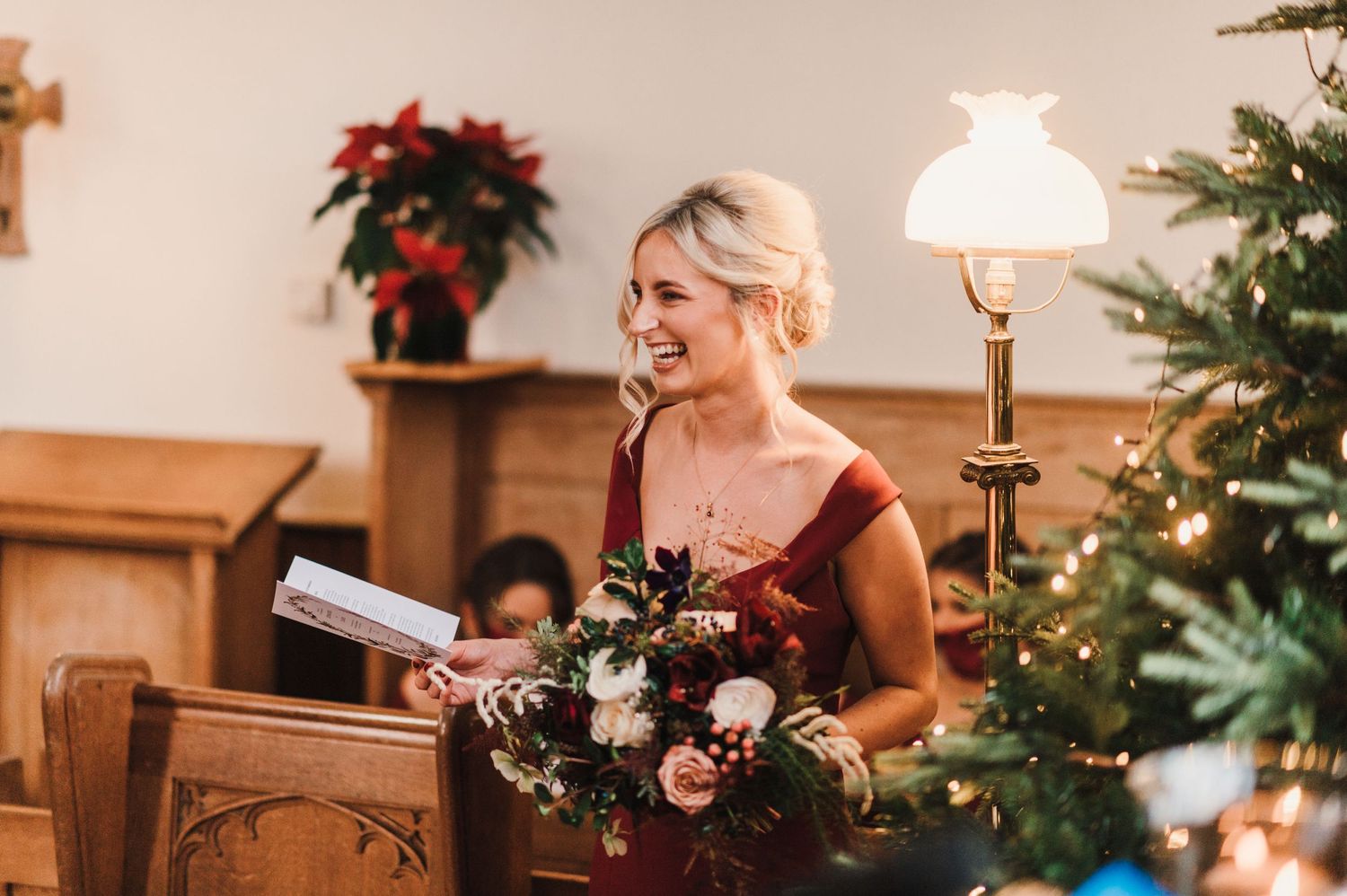 Wedding ceremony attendant holds bouquet inside traditional church decorated with poinsettias and Christmas decor.