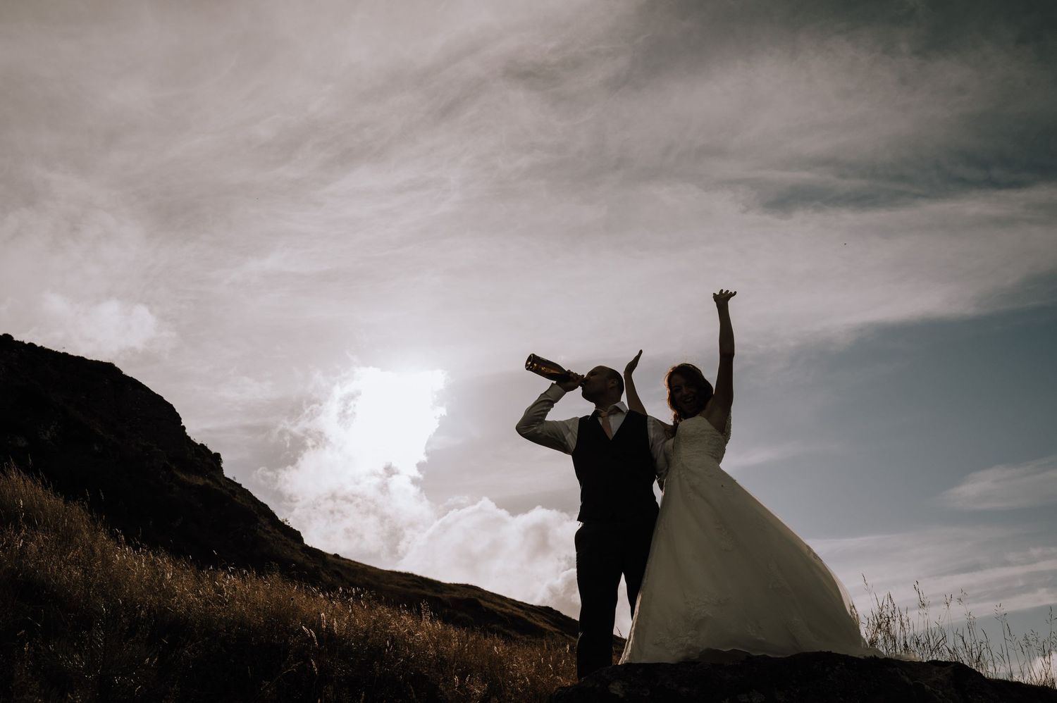 Two figures celebrate with champagne against a dramatic mountain sunset backdrop.