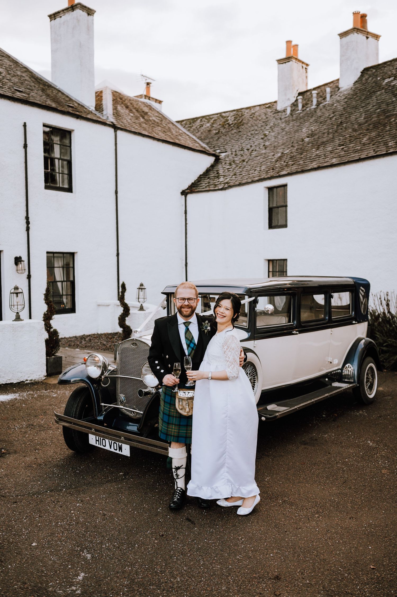 Wedding couple stands beside a classic vintage car against white-walled Scottish architecture.