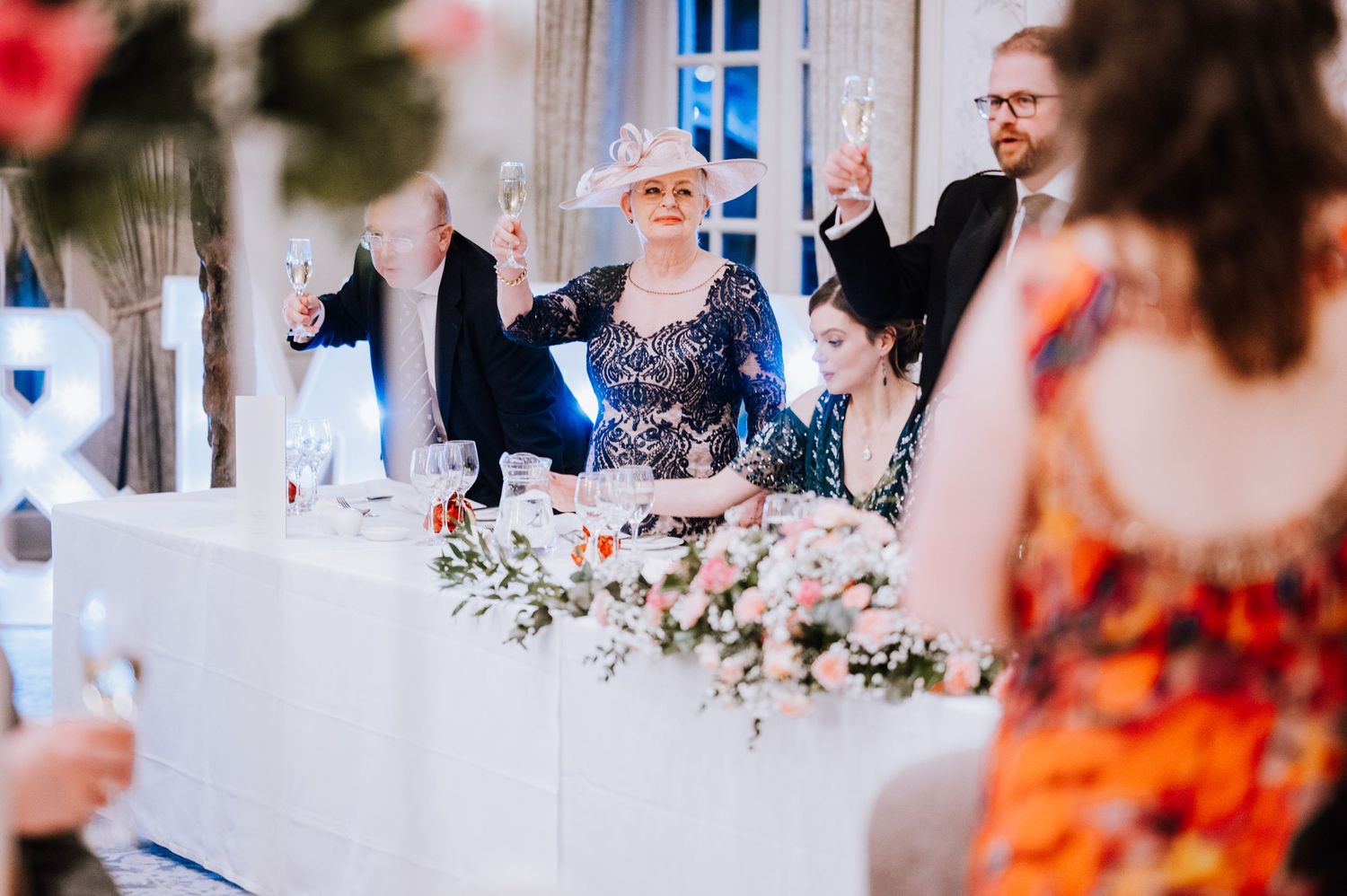 Guests at head table celebrating with raised arms during wedding reception.