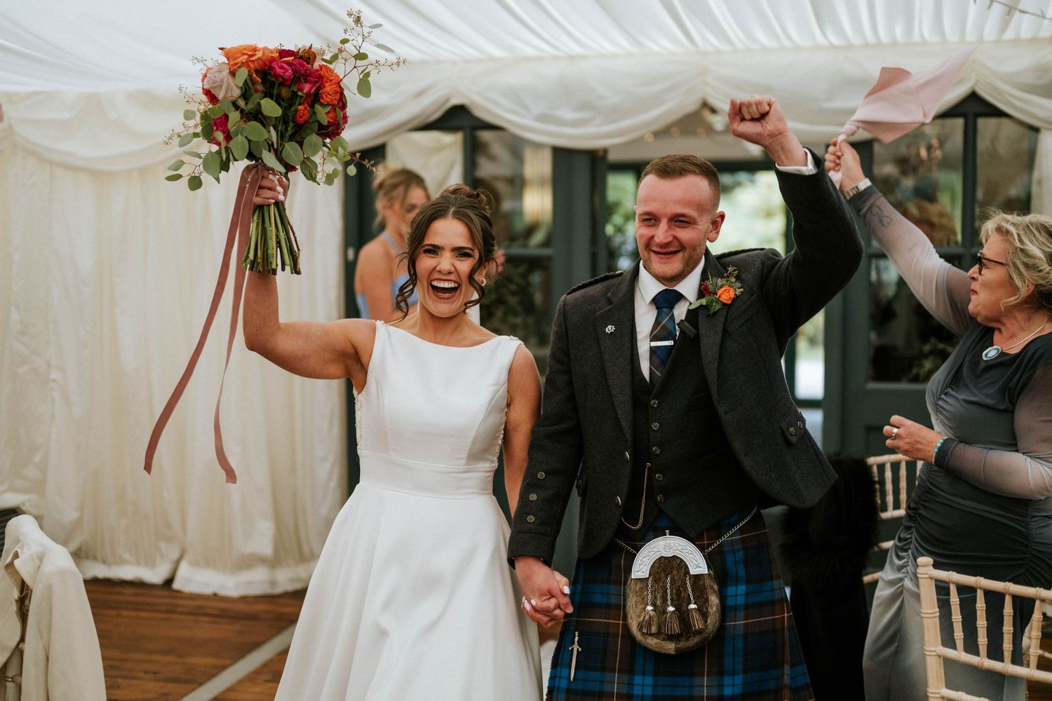 A celebration moment under a white marquee tent with Scottish wedding attire and floral decorations.