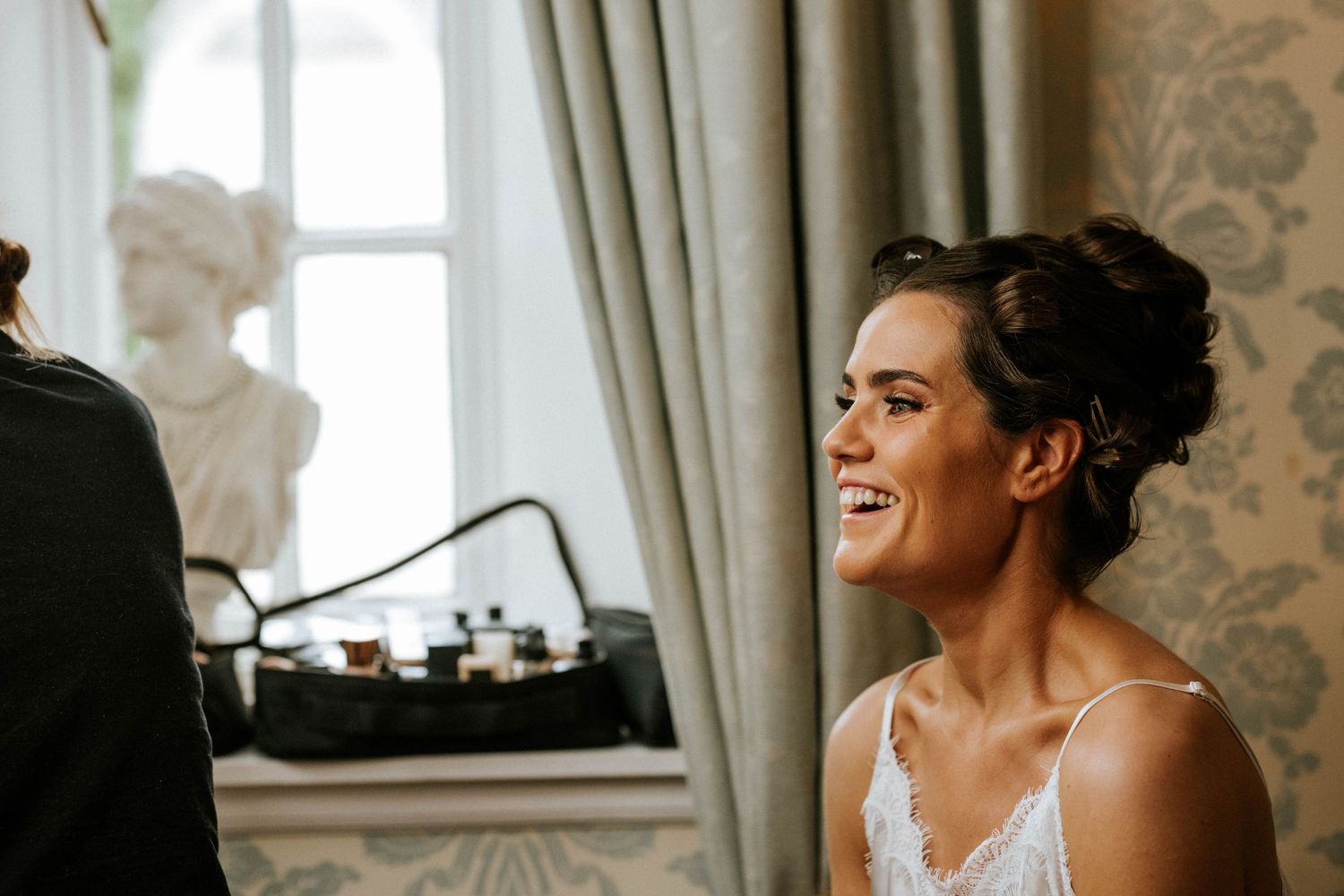 A bride in a white wedding dress stands by a window with natural light streaming in.
