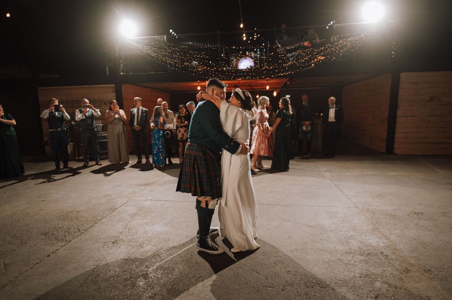 A couple shares their first dance at a Scottish wedding reception in a dimly lit venue with string lights overhead.