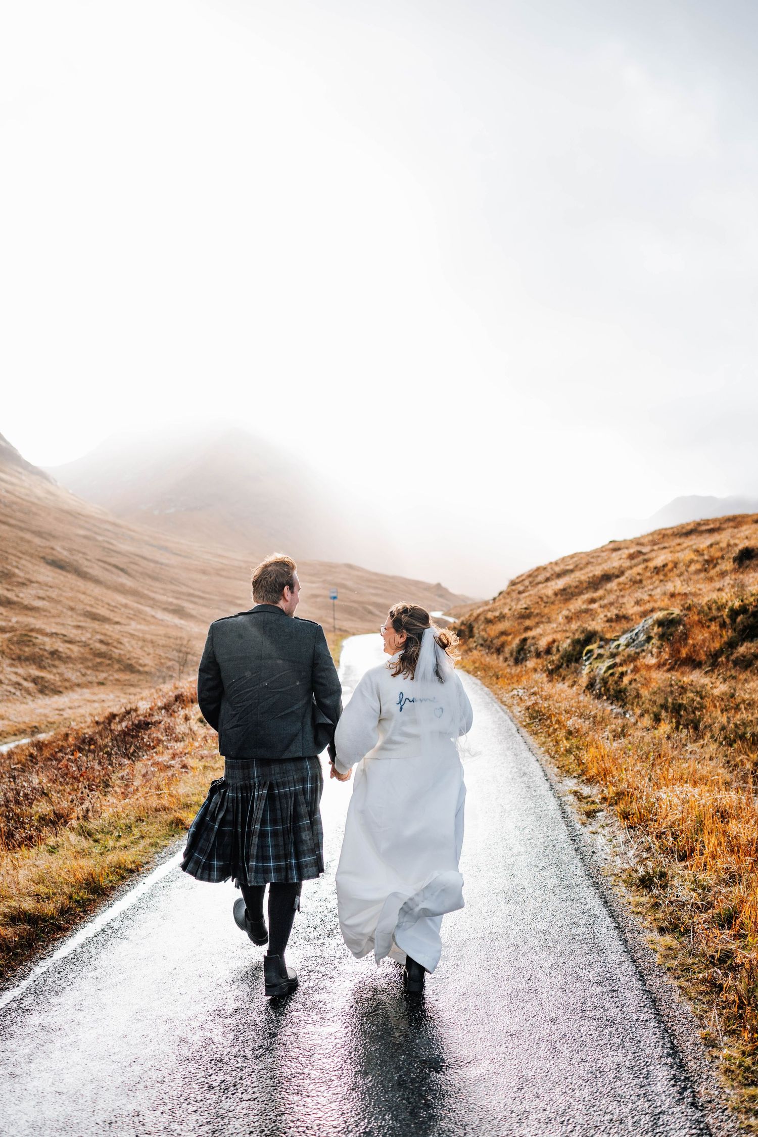 A couple walks down a misty road in the Scottish Highlands, with the groom in a kilt and bride in white dress.