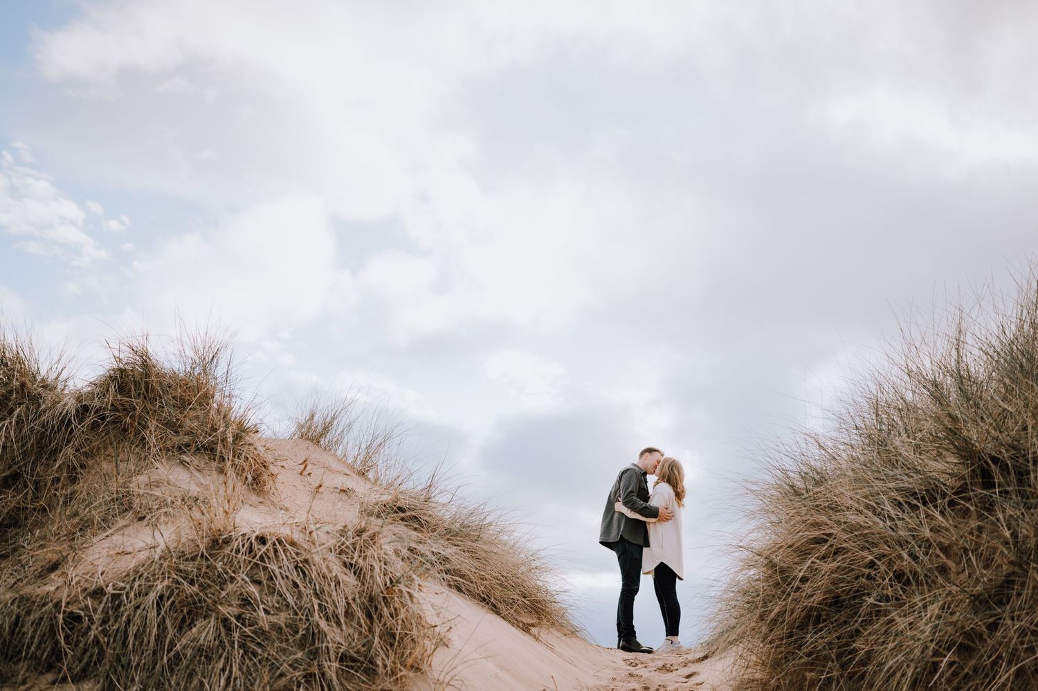Couple embraces among tall beach grass on coastal dunes at sunset.