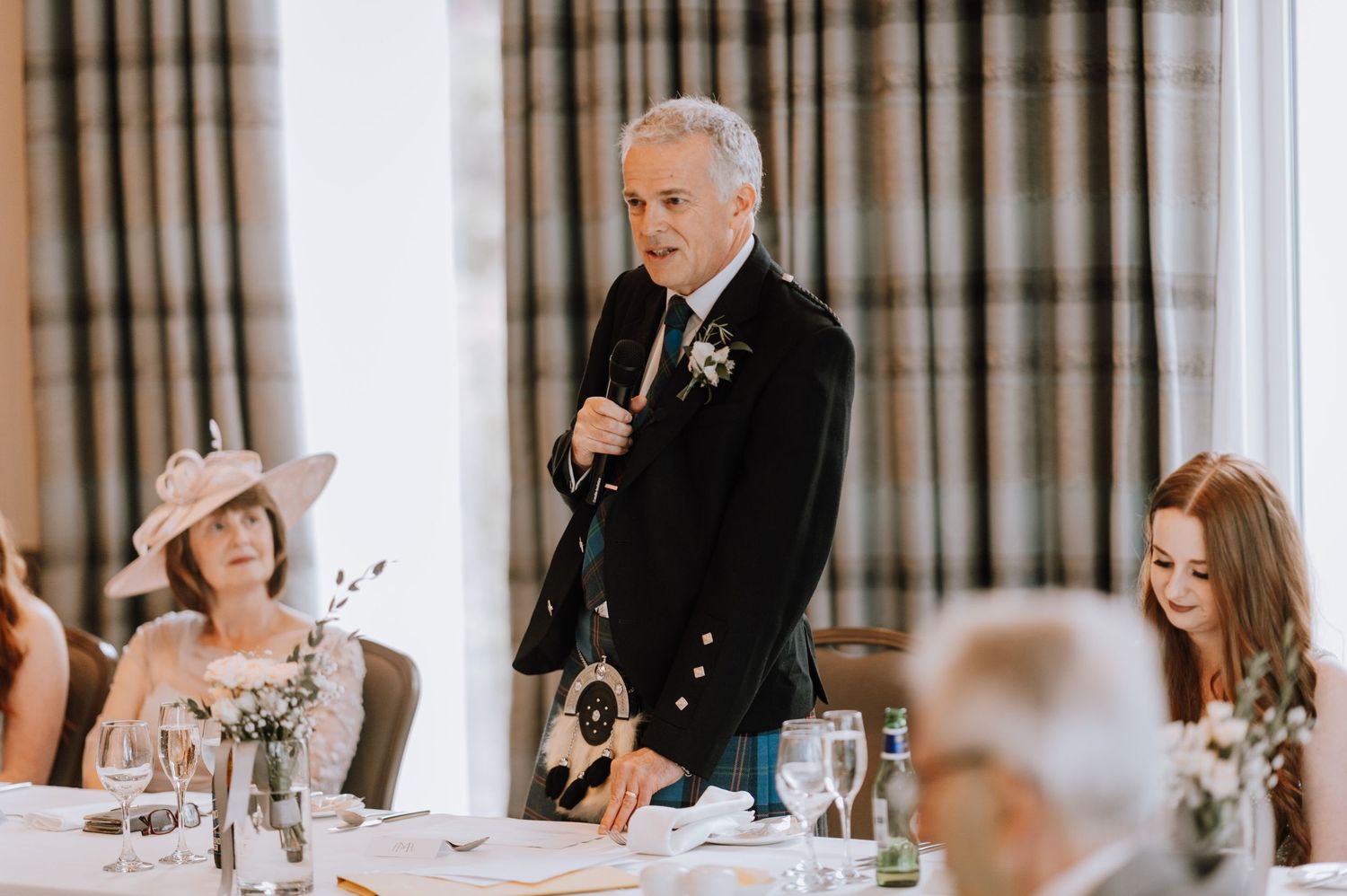 A speaker in traditional Scottish formal wear addressing wedding guests during dinner.