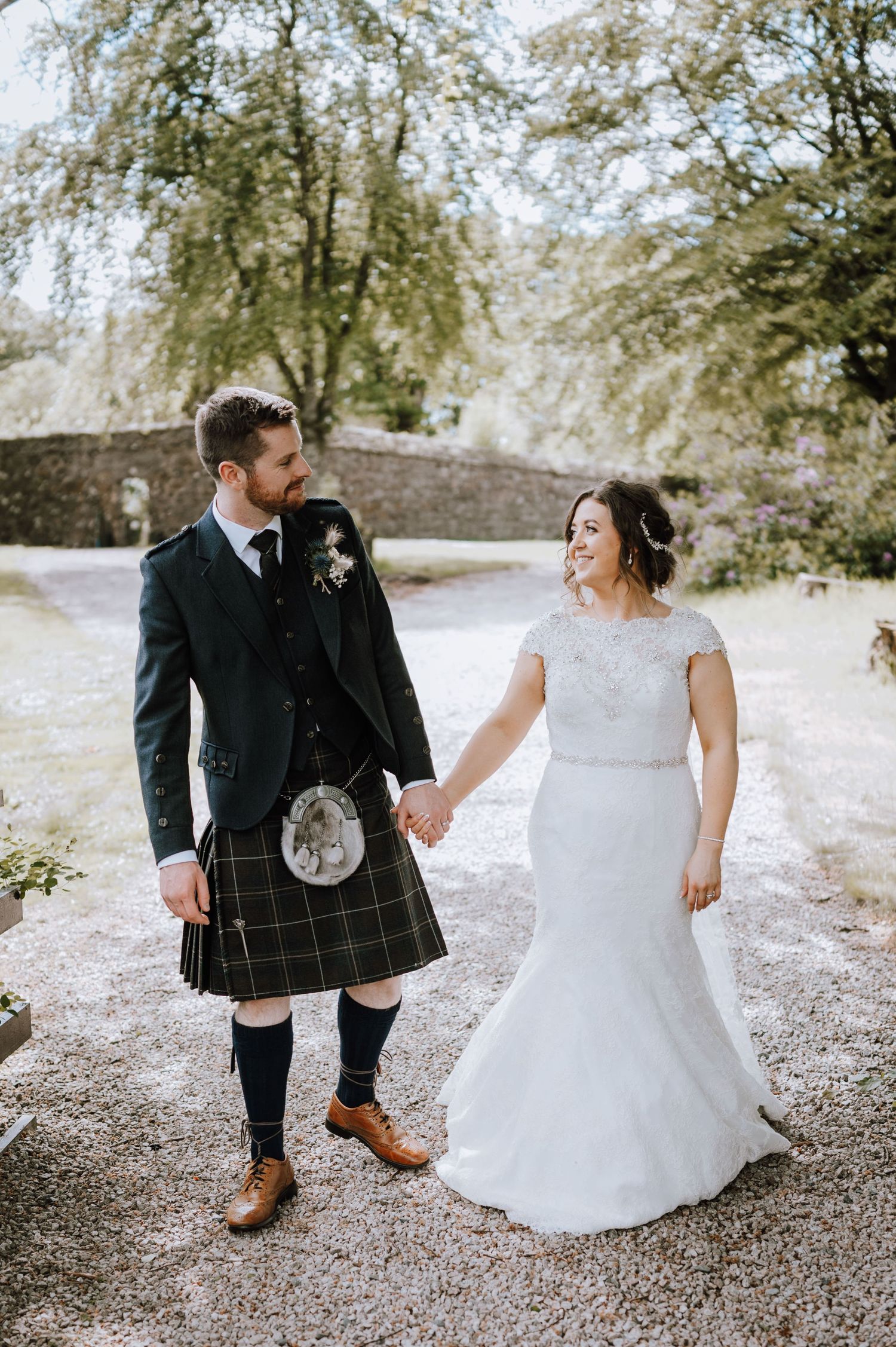 Bride and groom walking hand in hand on a path, showcasing traditional Scottish wedding attire.