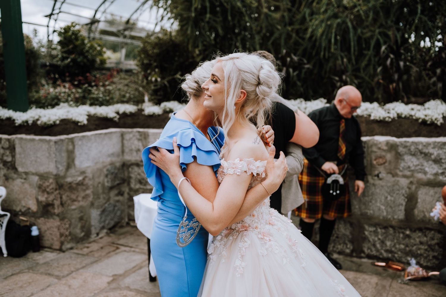 A tender embrace between wedding guests in blue and white dresses at an outdoor garden reception.