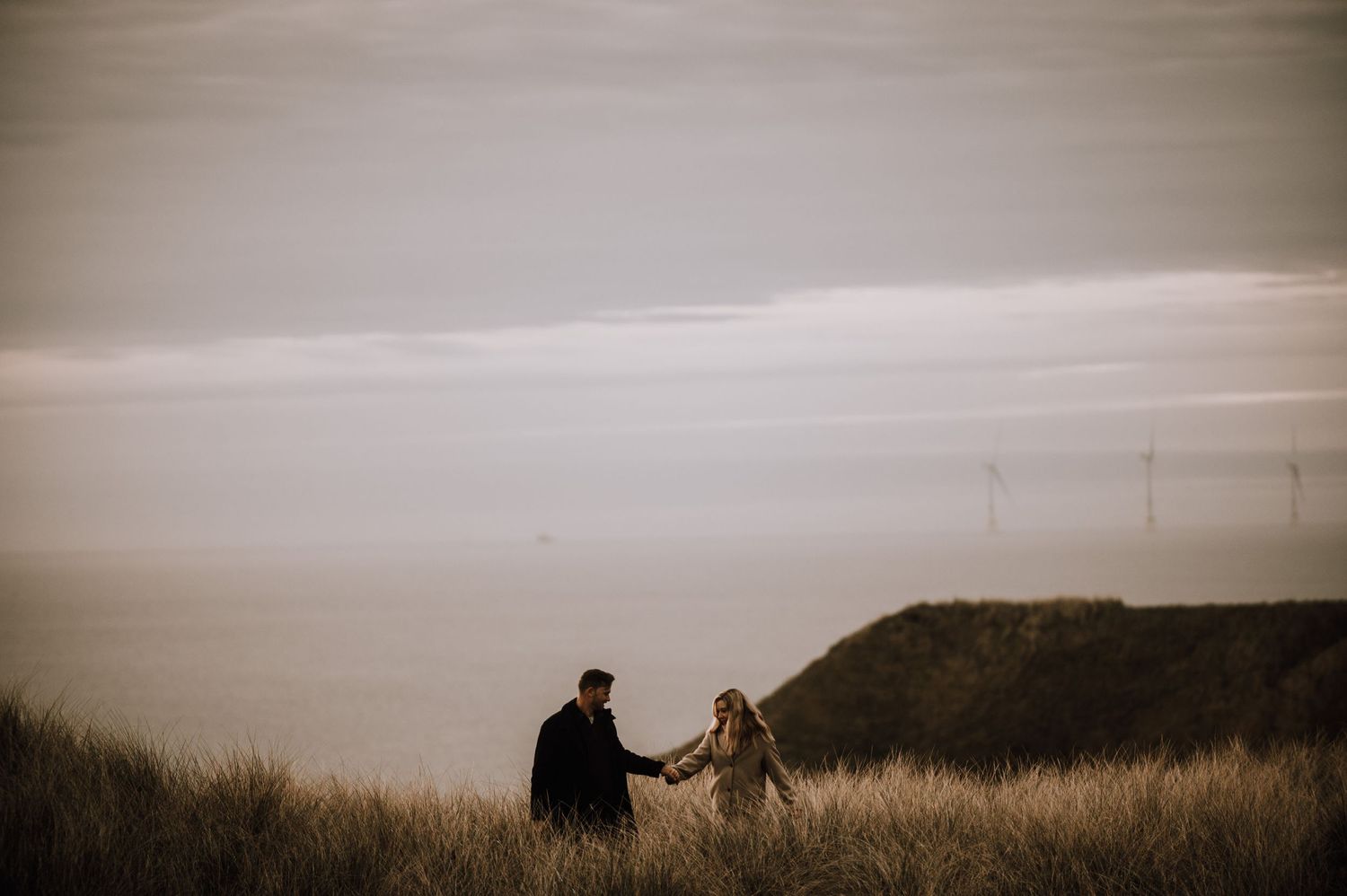 Two figures hold hands while standing on a misty coastal hillside.