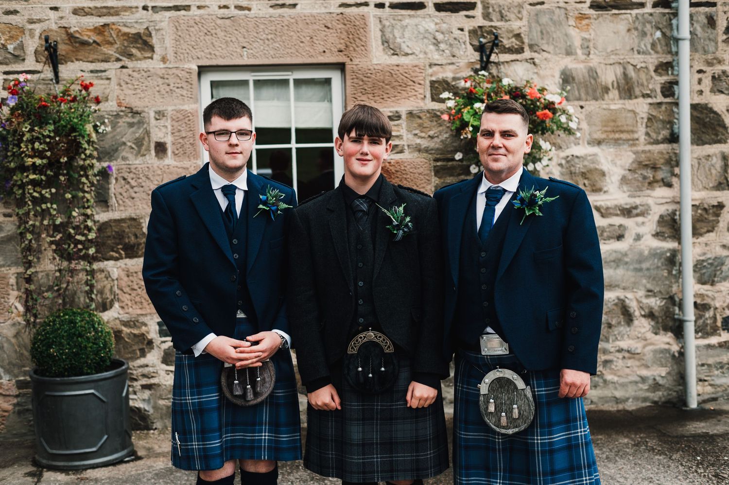 Groomsmen in traditional Scottish kilts and dark jackets pose together against stone wall backdrop.
