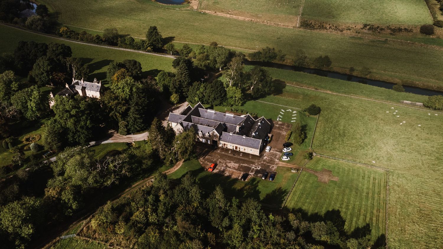 Aerial view of a rural estate with stone buildings surrounded by green fields and woodlands in the countryside.