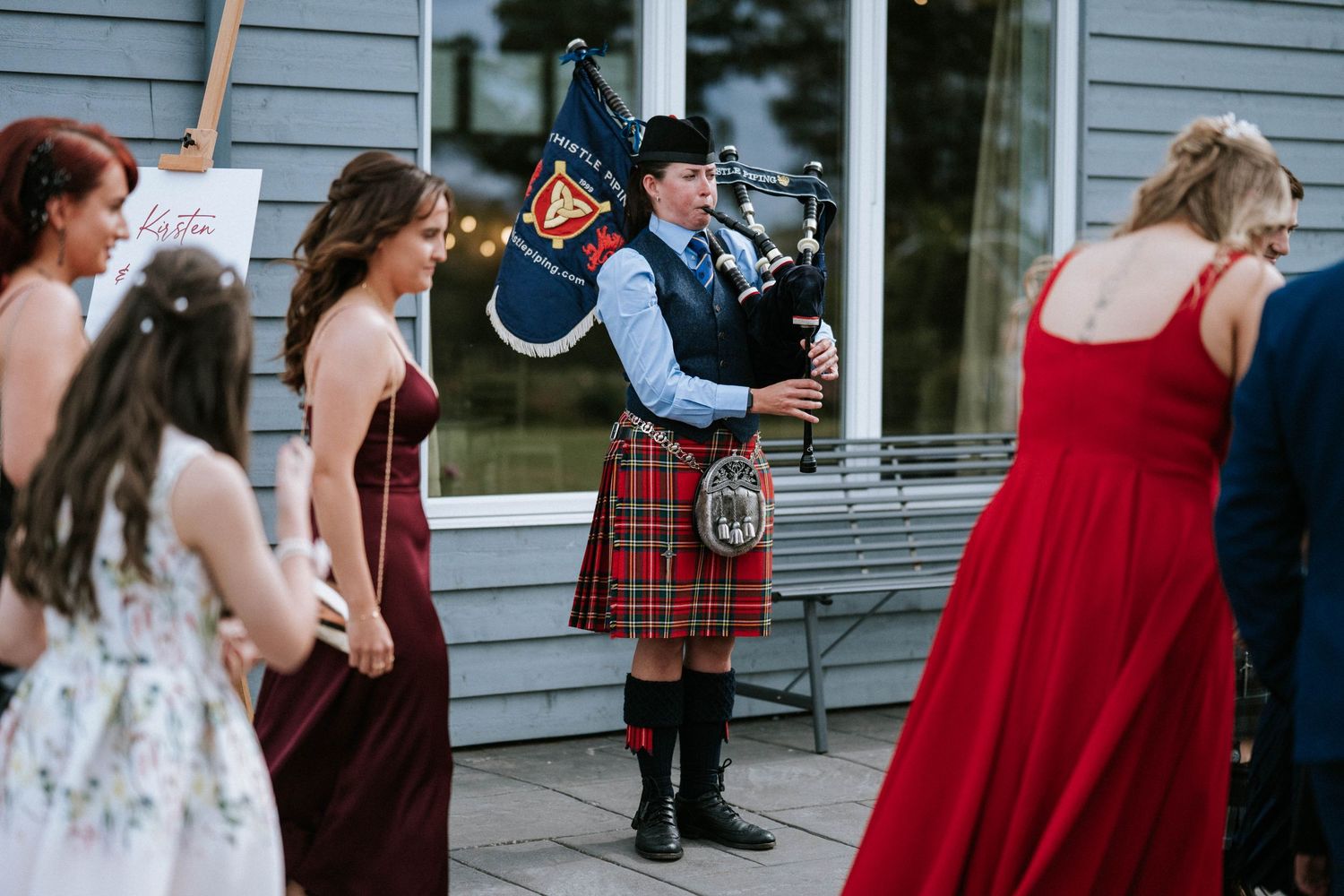 A bagpiper in traditional Scottish attire performs for guests in formal wear at an outdoor event.