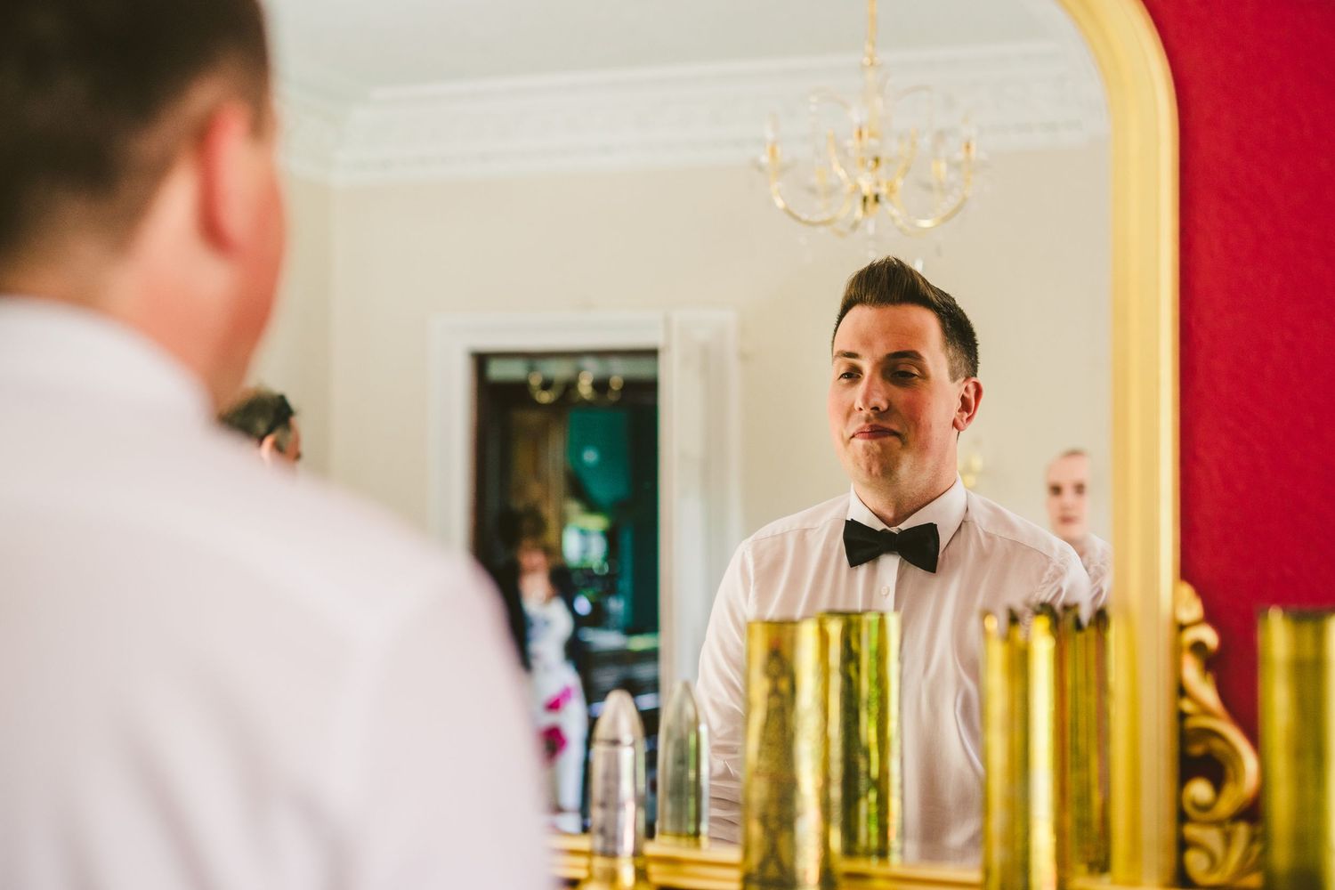 A person in a bow tie looks at their reflection in a mirror with gold decor.