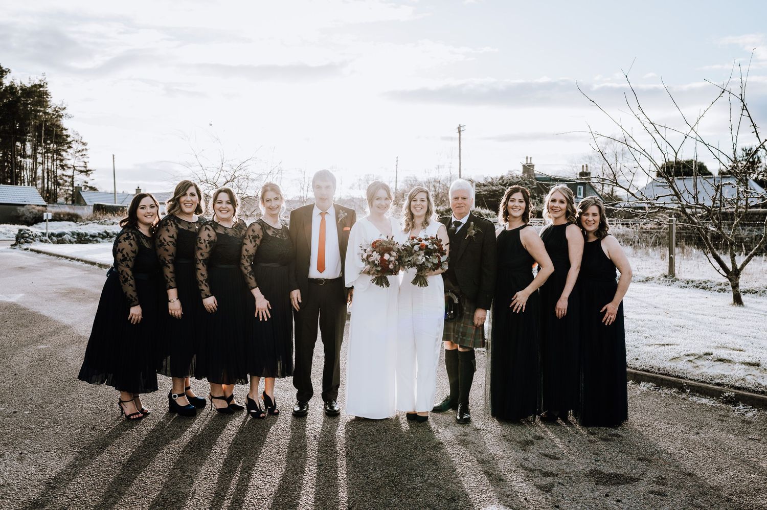 Same Sex Wedding Photography. Wedding party stands in line against winter landscape wearing black and white formal attire.
