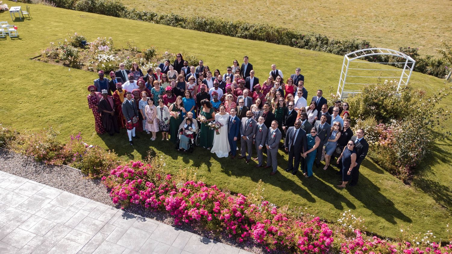 Large group of wedding guests gathered on a sunny hillside with colorful flowers in the foreground.