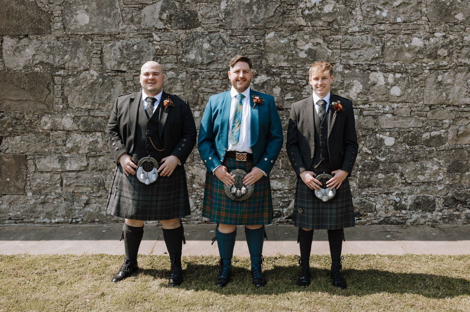 Three people in traditional Scottish kilts and formal wear standing against stone wall.