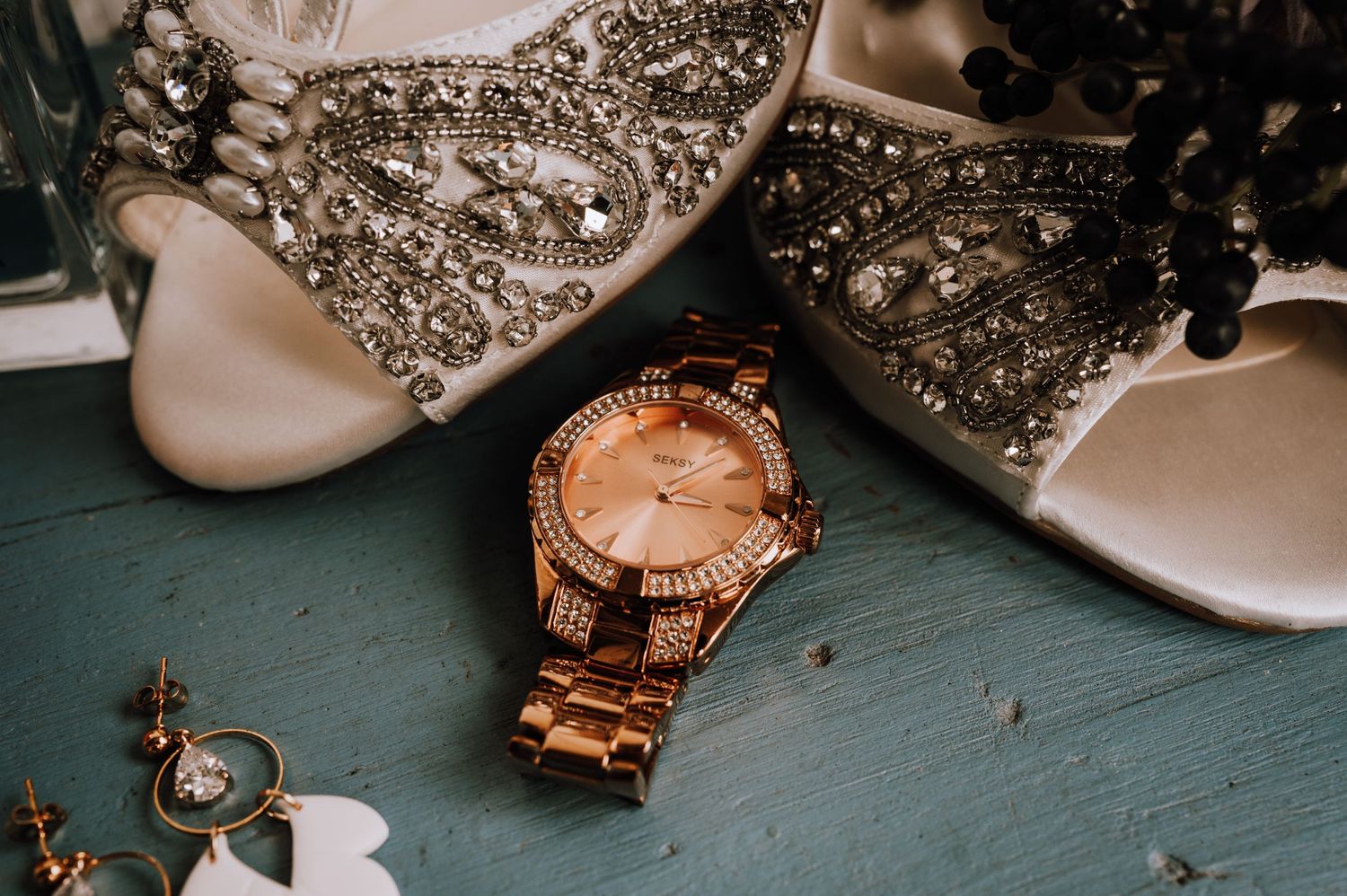 Rose gold watch and jeweled wedding shoes arranged on turquoise surface.